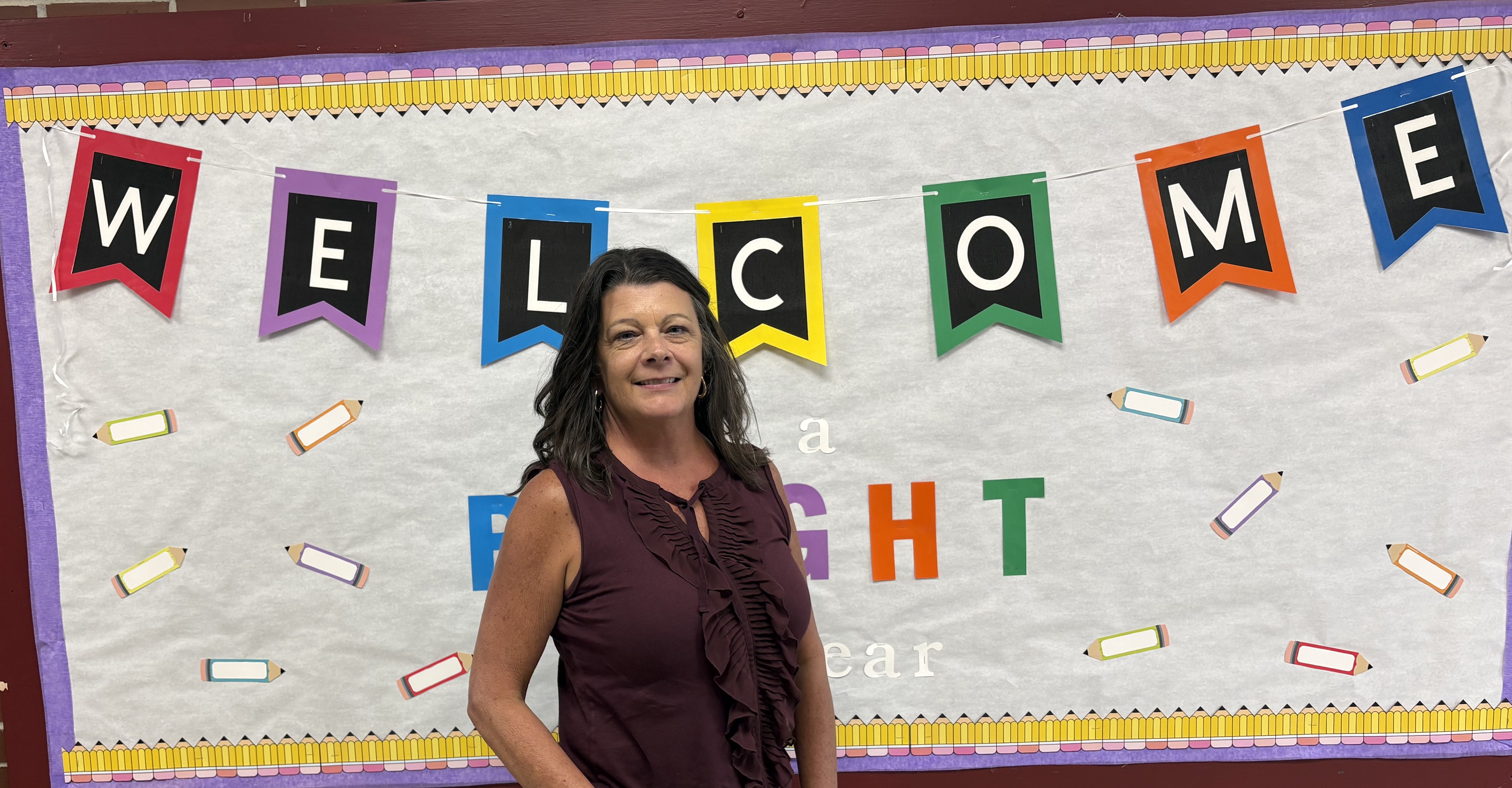 A woman in a maroon top stands in the foreground against a background of a "WELCOME" banner and school-themed decorations.
