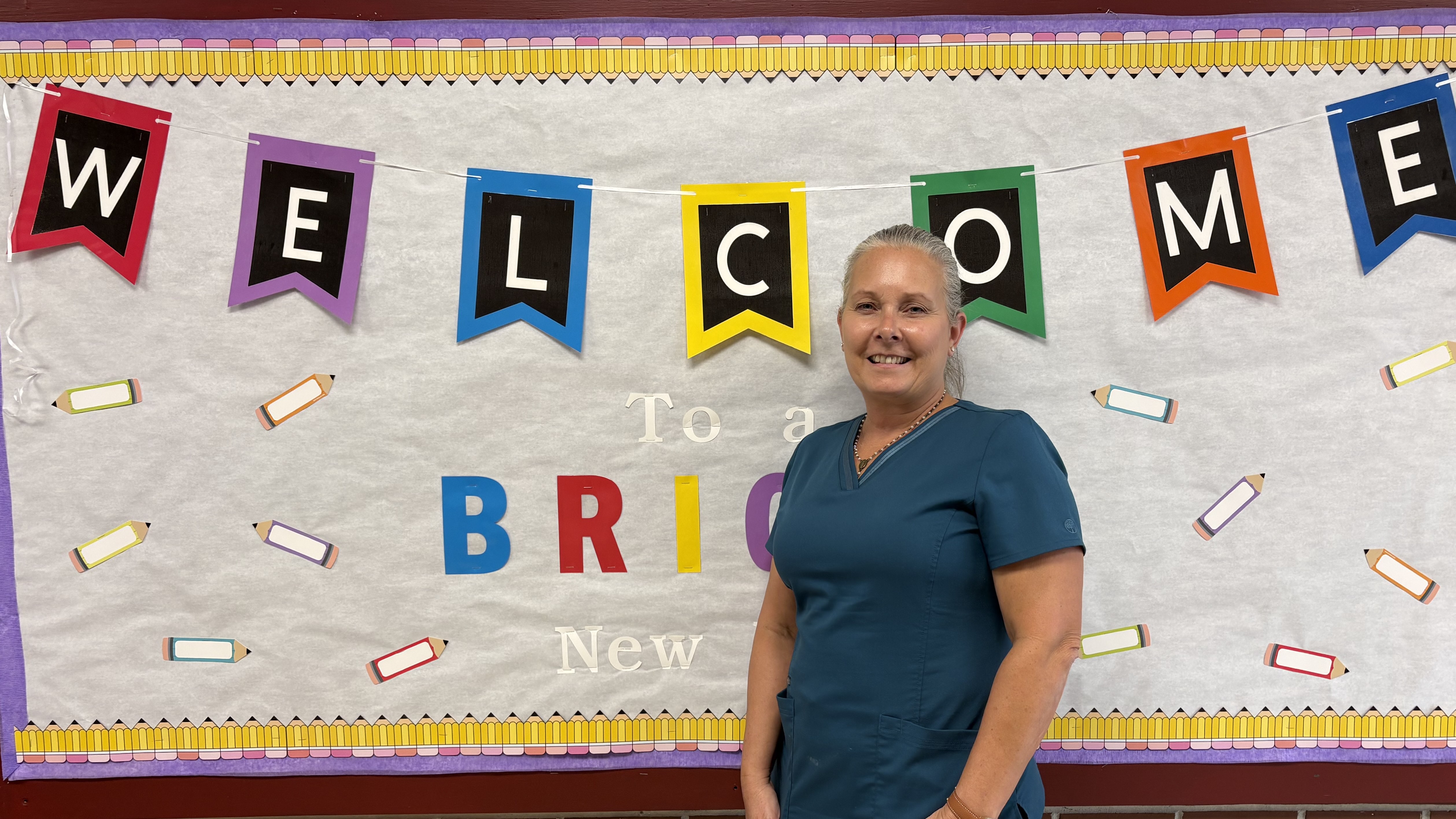 A woman in blue scrubs stands in the foreground before a white banner with colorful "WELCOME" flags and pencil illustrations in the background.