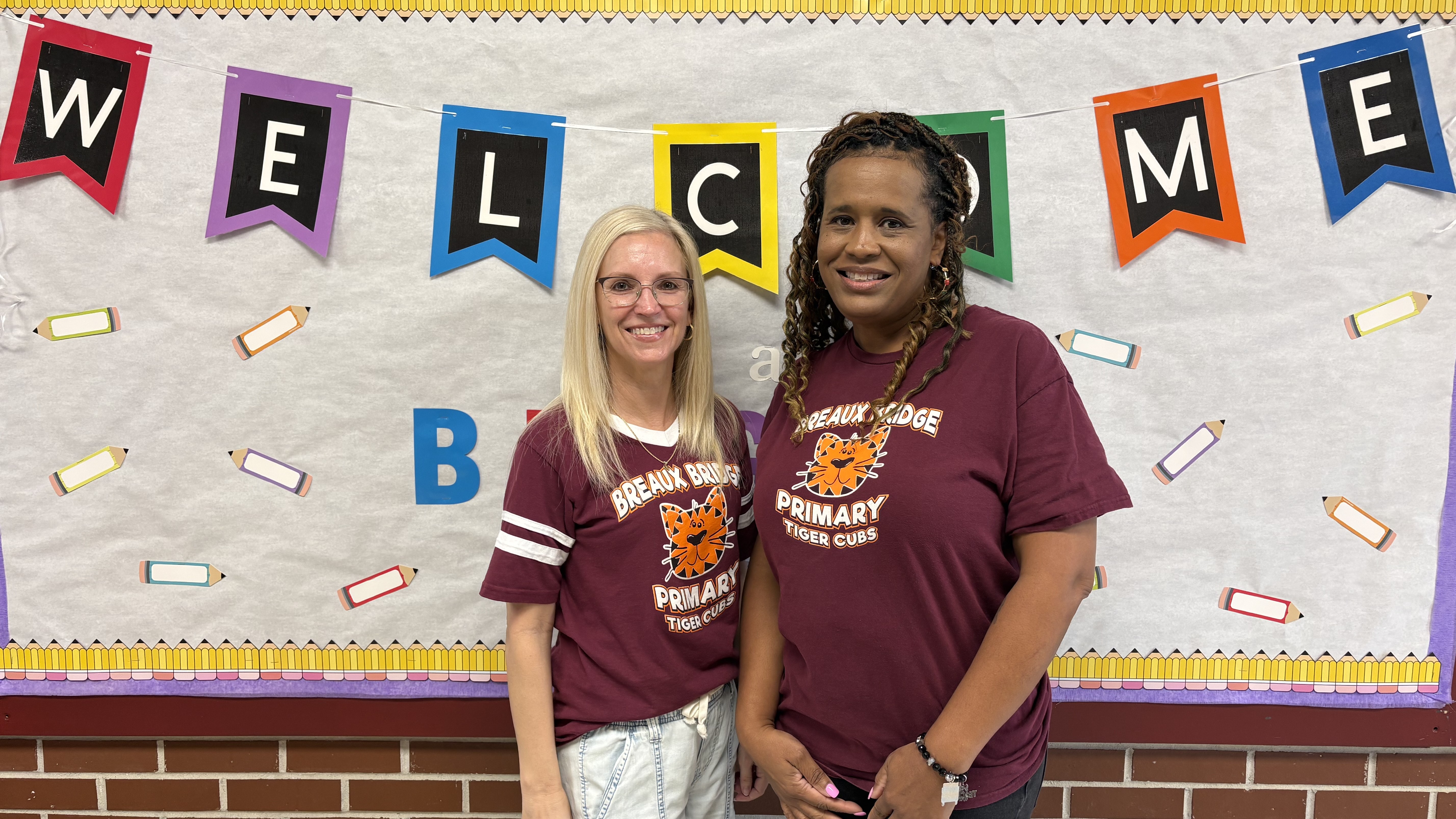 Two women wearing maroon t-shirts stand in front of a white bulletin board decorated with a colorful "WELCOME" banner and pencil illustrations.