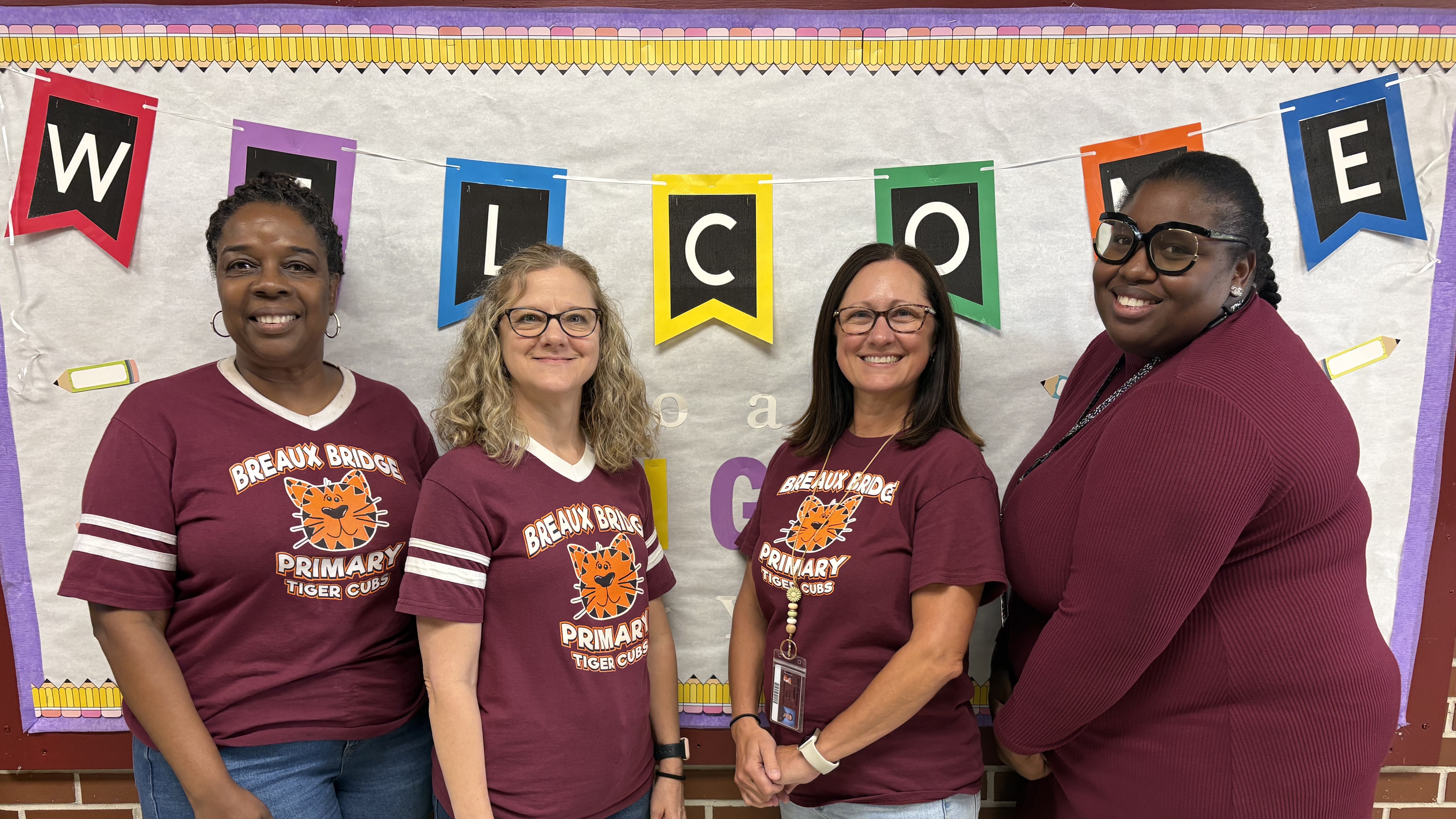 Four women stand smiling in the foreground against a classroom background featuring a "WELCOME" banner.