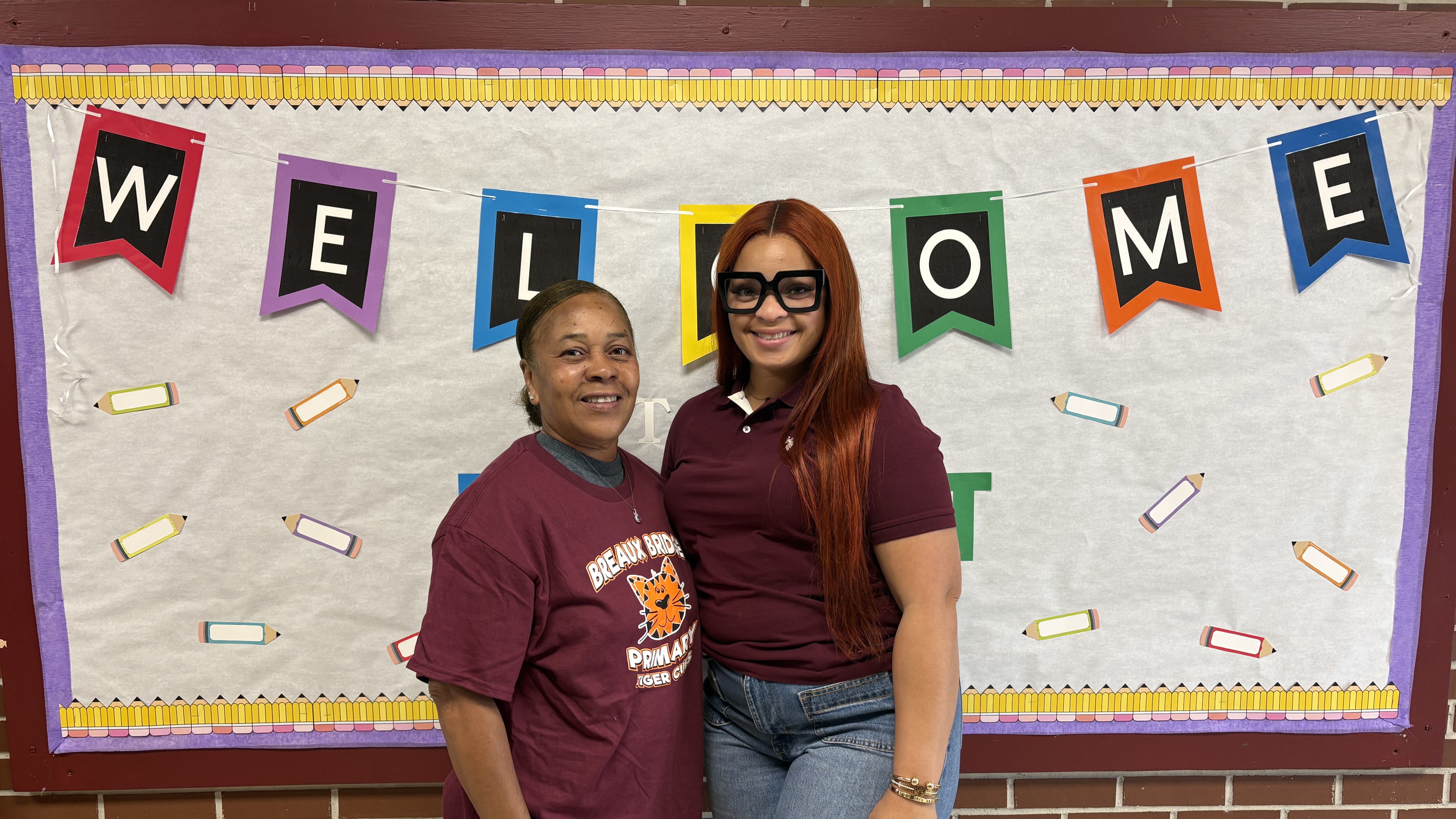 Two women stand side-by-side in the foreground in front of a "WELCOME" banner decorated with pencils.