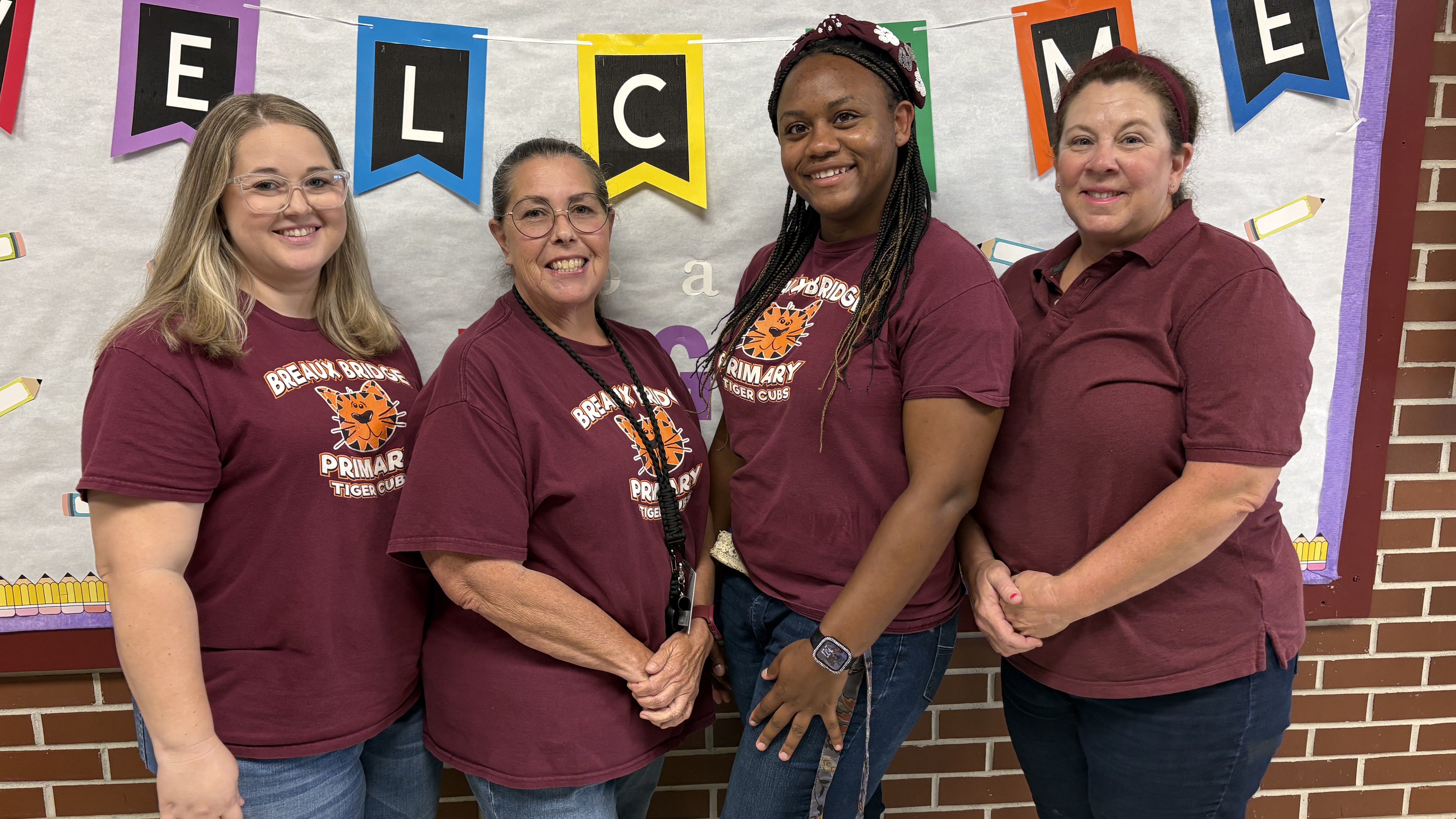 Four women wearing maroon shirts stand together in front of a "WELCOME" banner and a brick wall.