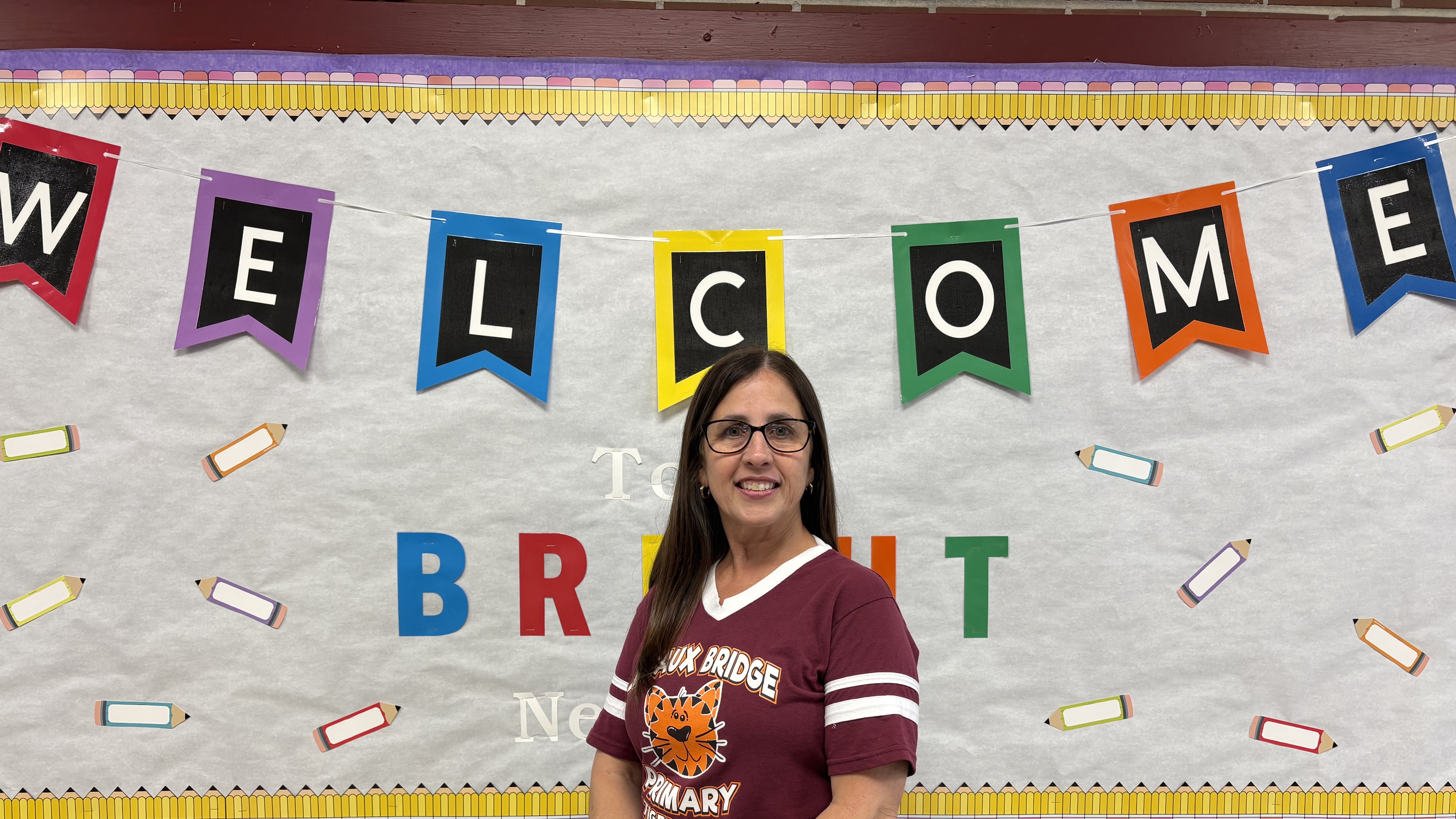 A smiling woman in a maroon shirt stands in front of a white backdrop decorated with a colorful "WELCOME" banner and pencil illustrations.
