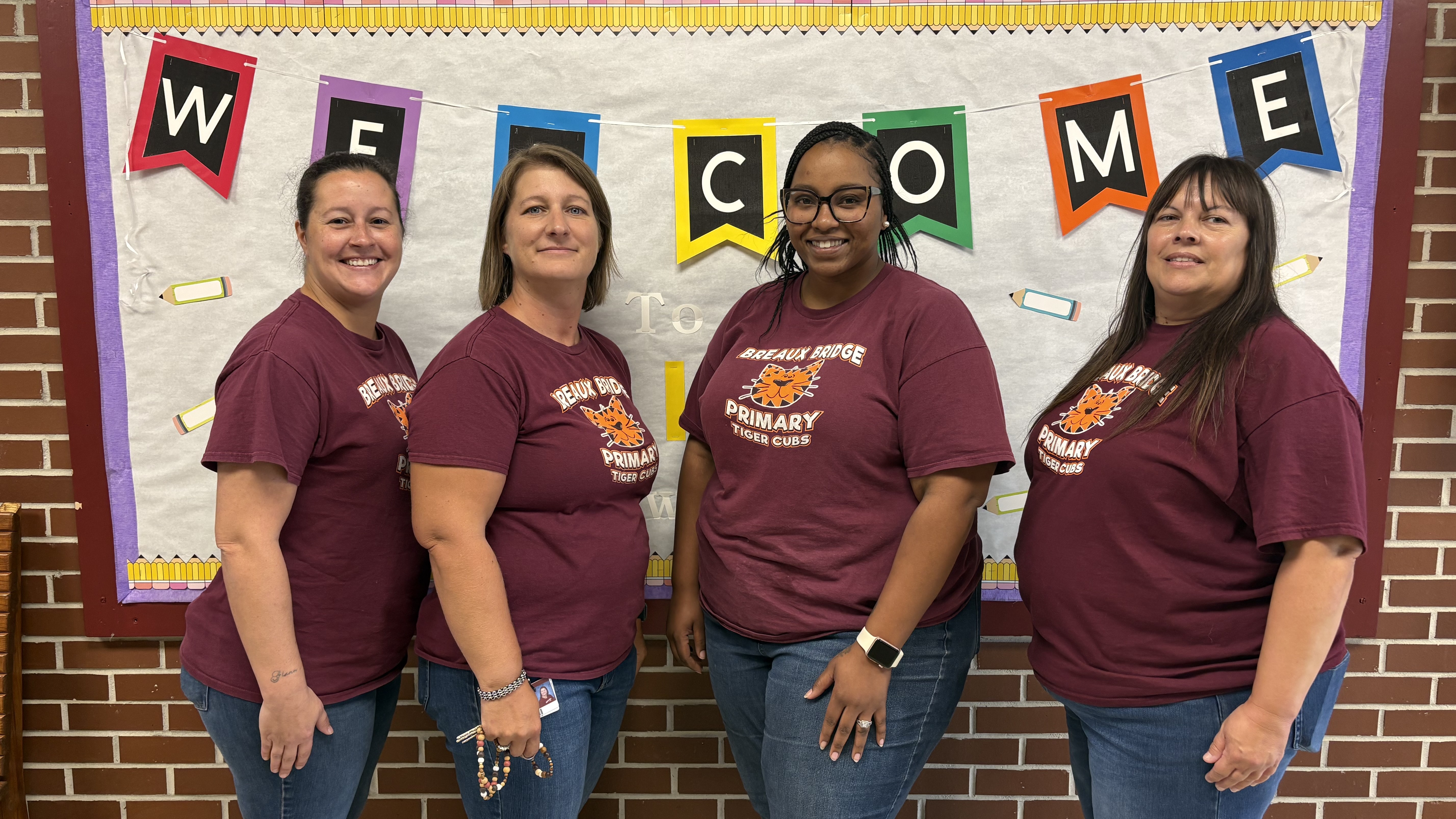Four women wearing matching maroon t-shirts stand in the foreground in front of a "WELCOME" banner and a brick wall.