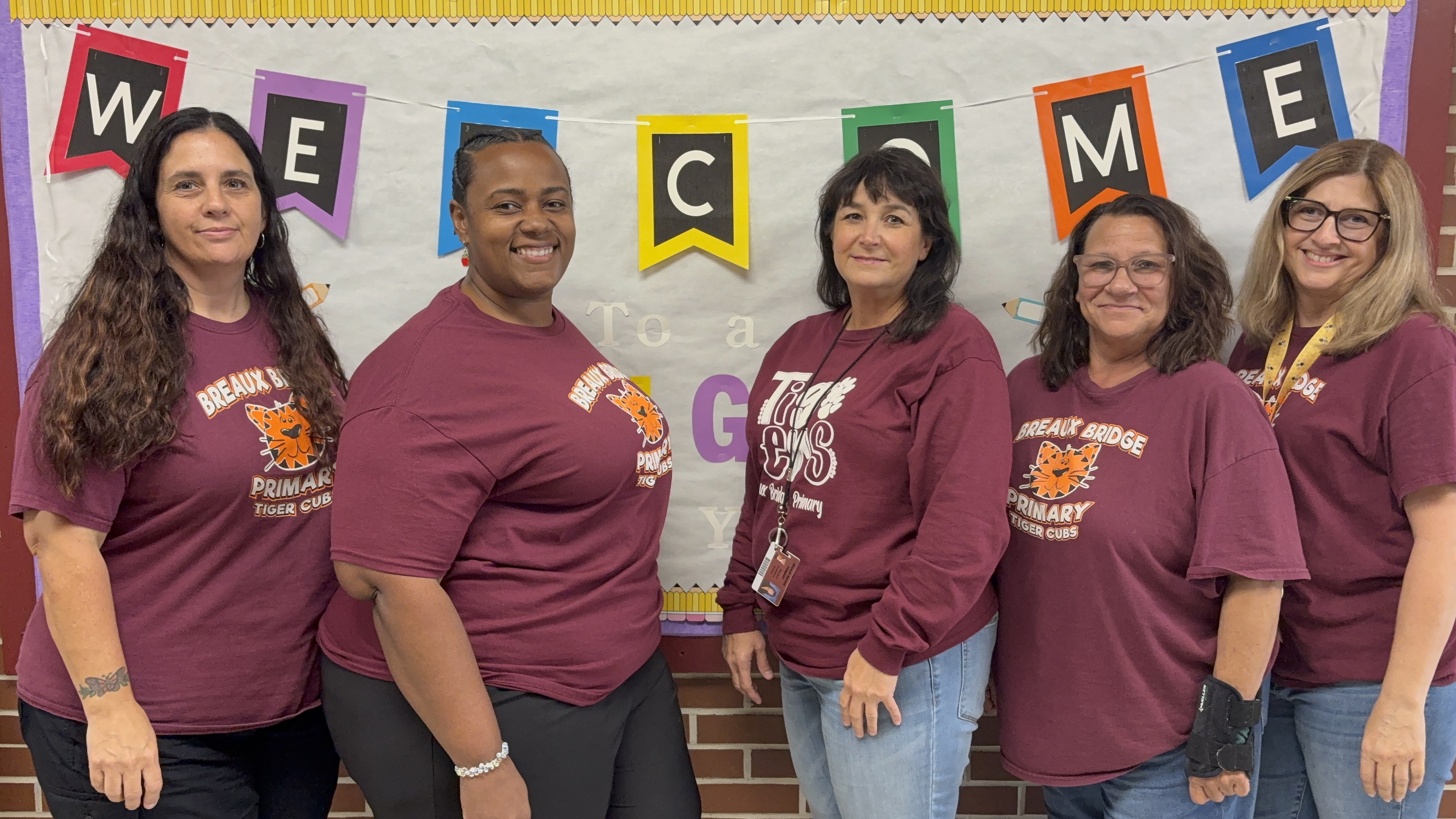 Five women wearing maroon shirts stand in the foreground in front of a "WELCOME" banner and a brick wall.