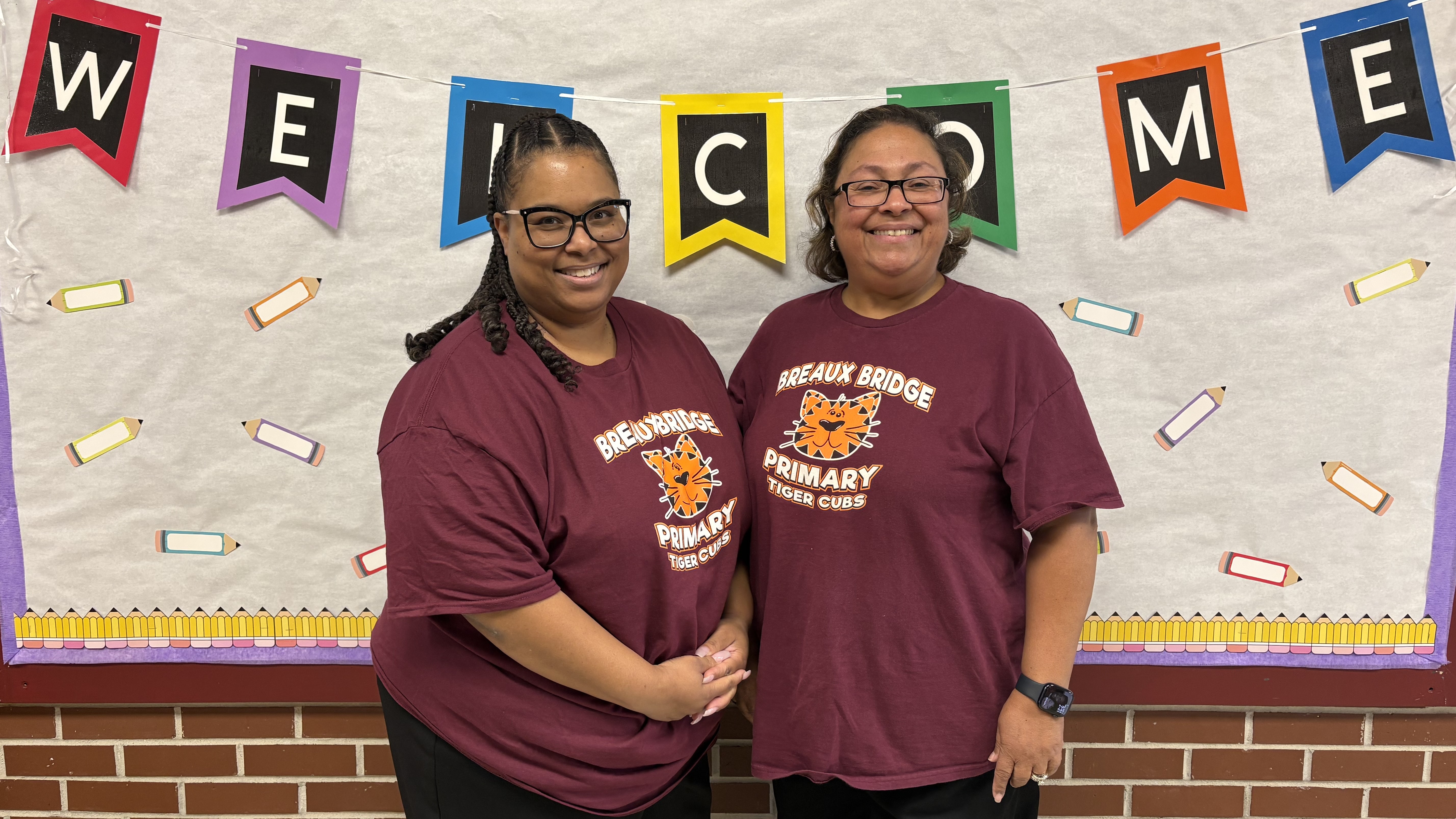 Two women wearing maroon t-shirts stand in the foreground against a background featuring a "WELCOME" banner and pencil decorations.