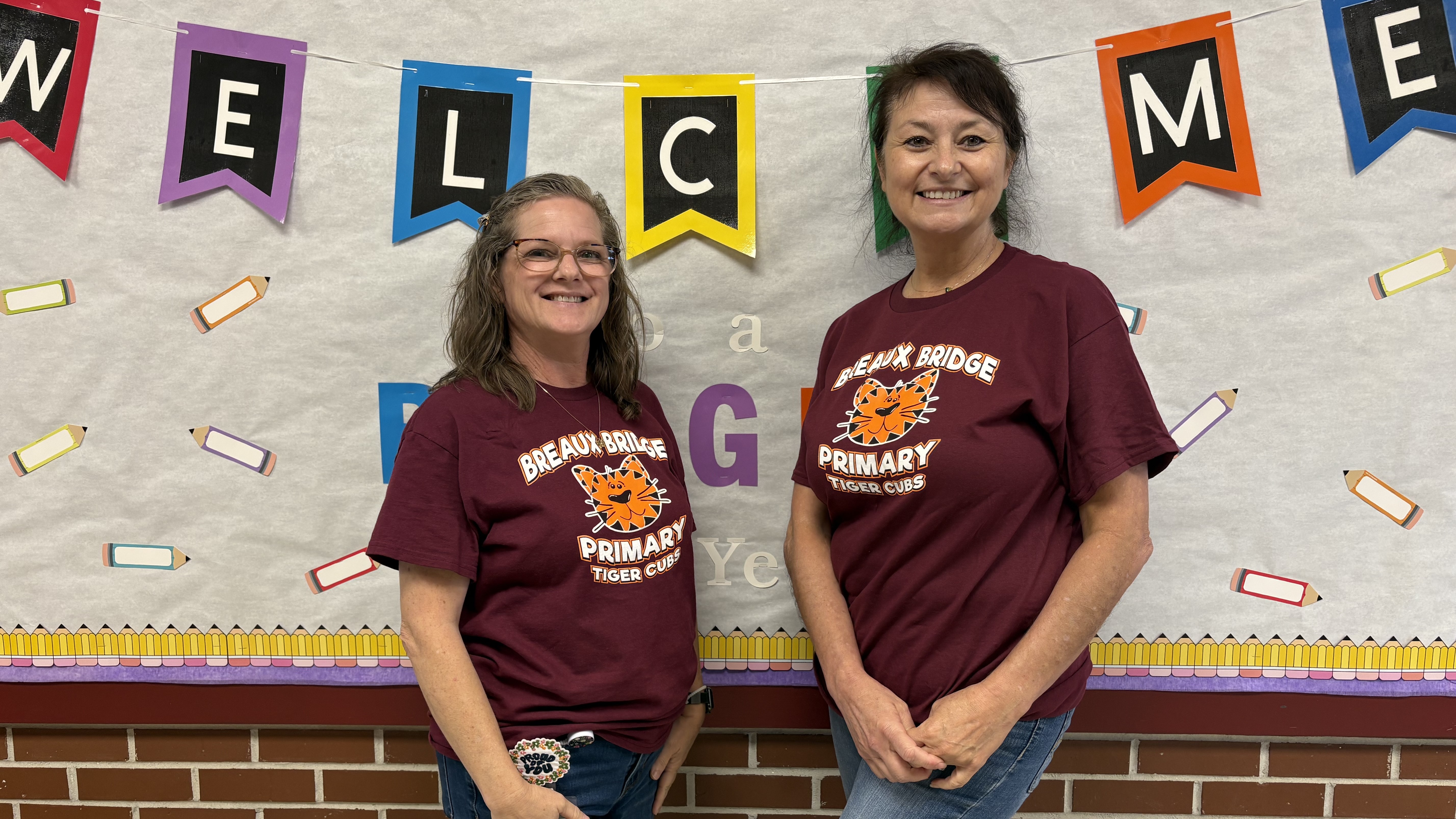 Two smiling women in matching maroon shirts stand in front of a bulletin board decorated with a "WELCOME" banner and colorful drawings, with a brick wall visible at the bottom.