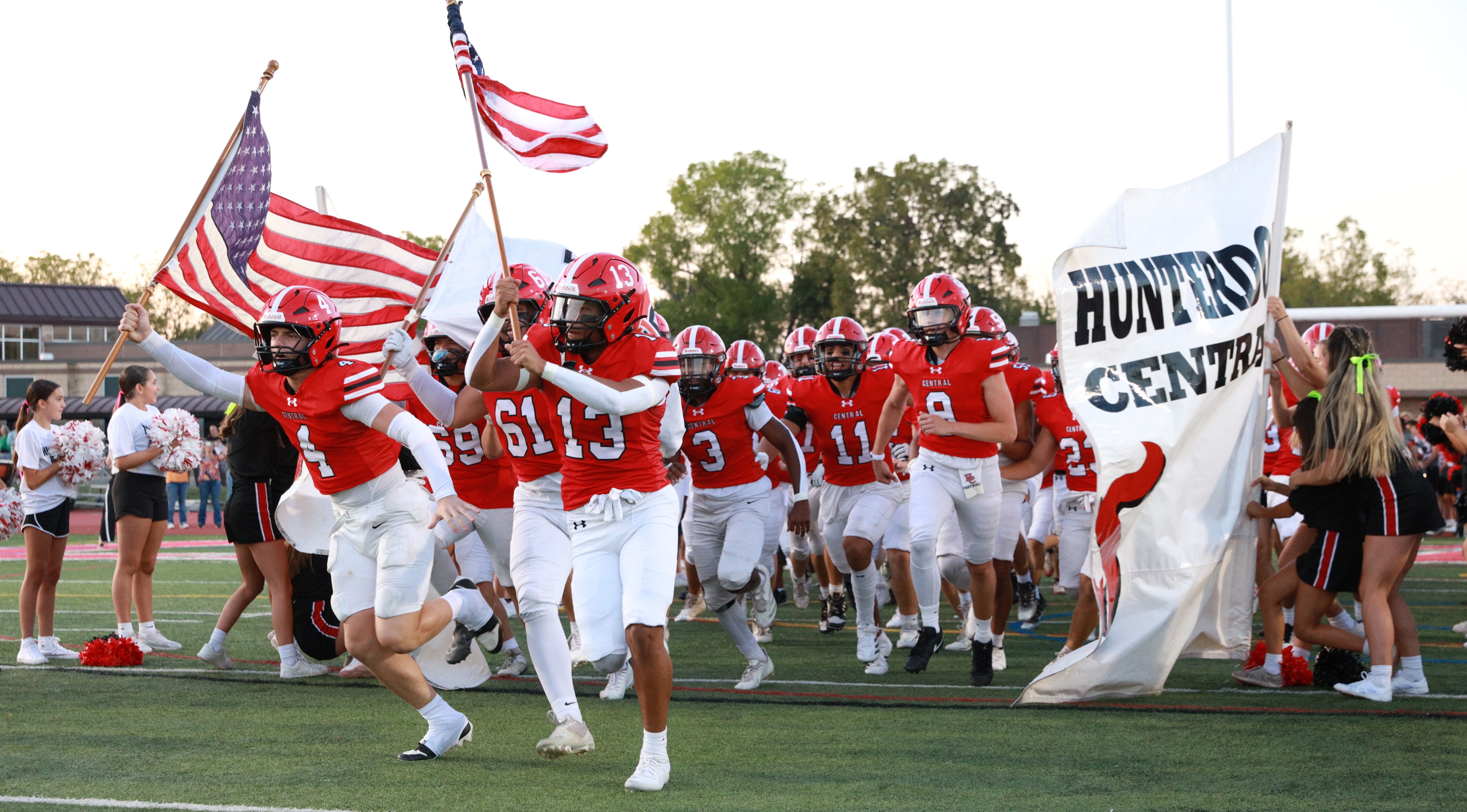 football players in red and white uniforms carry american flags and run through banner