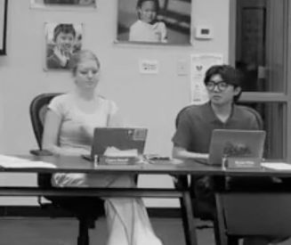 Black and white photo where male and female student sit at table in front of a large screen