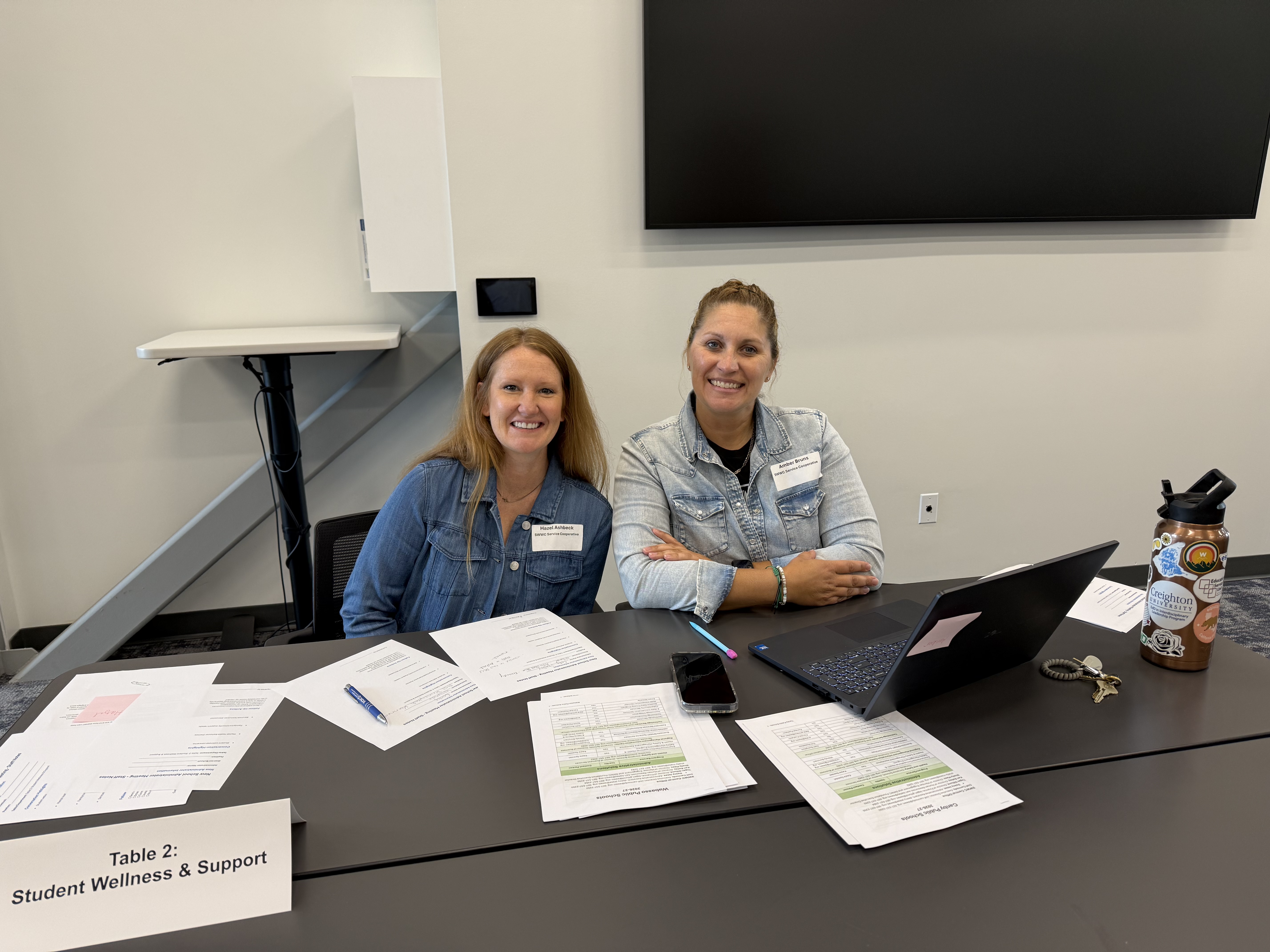 Two smiling women sit behind a table with papers and a laptop, with a large screen visible in the background.