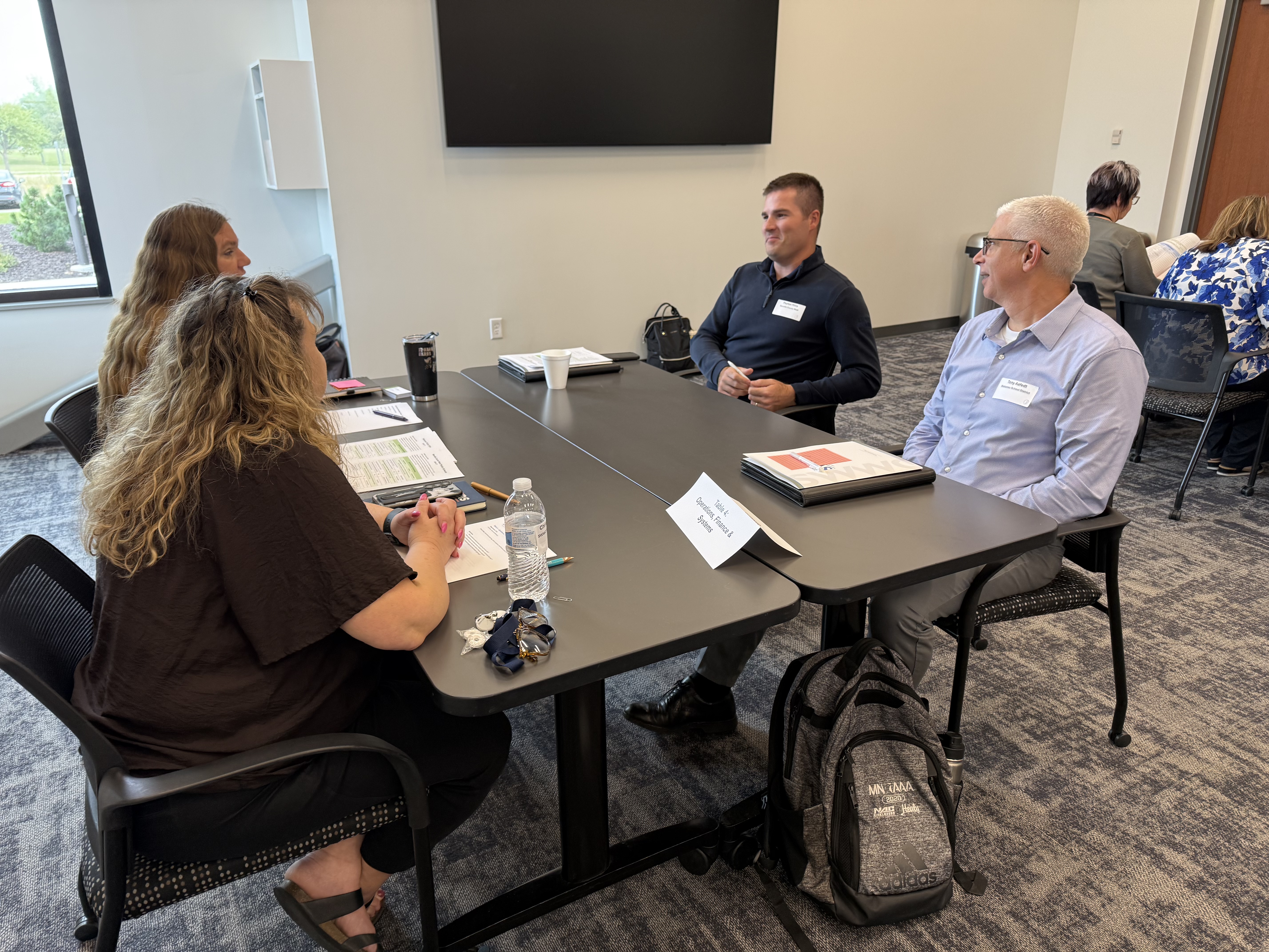 Four people sit around a conference table in the foreground, while a large screen and a doorway are visible in the background.