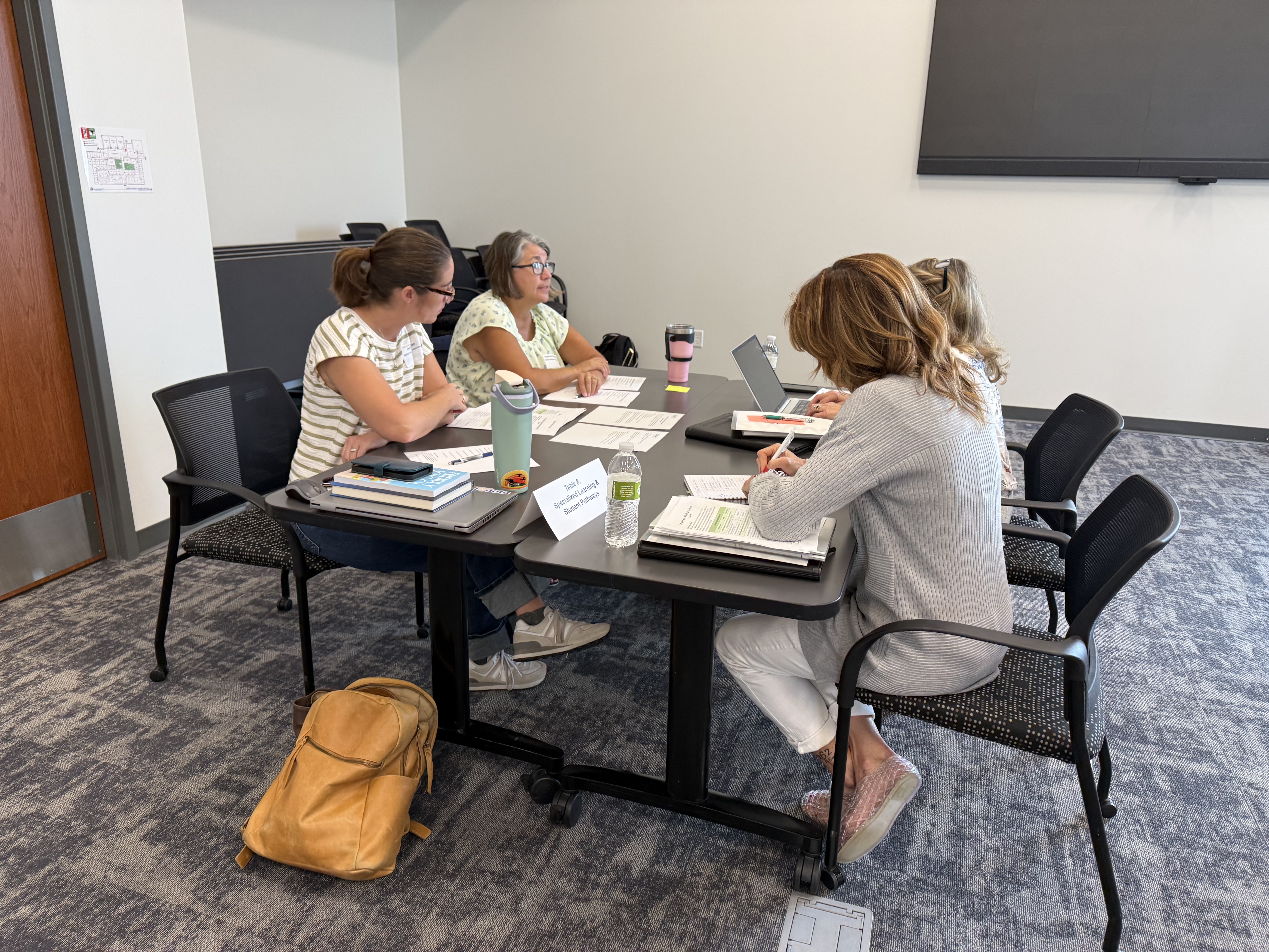 Four people sit around a table with papers and laptops in a brightly lit room with a projector screen on the back wall.