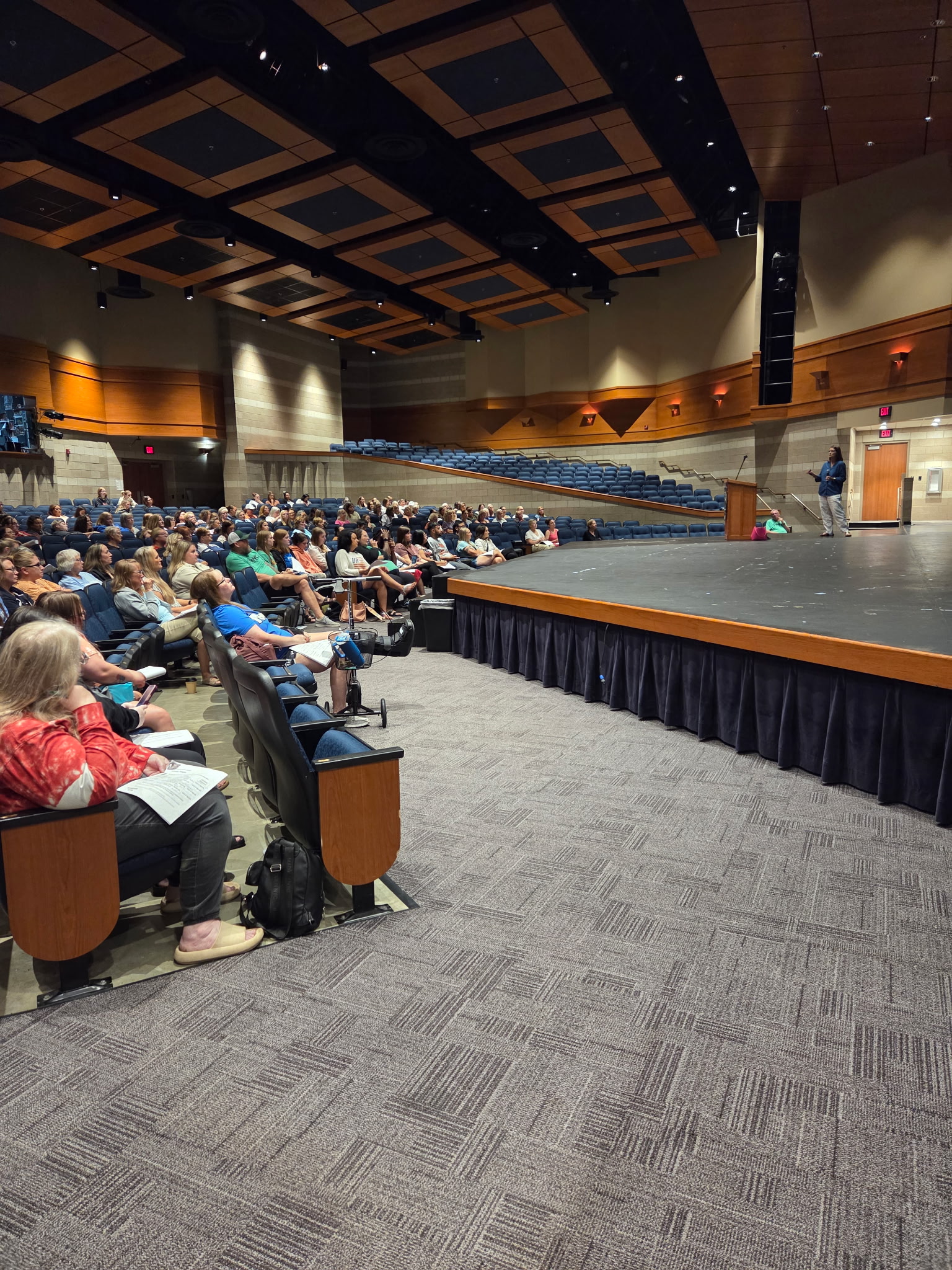 A speaker addresses a seated audience in a large auditorium with tiered blue seating in the background.