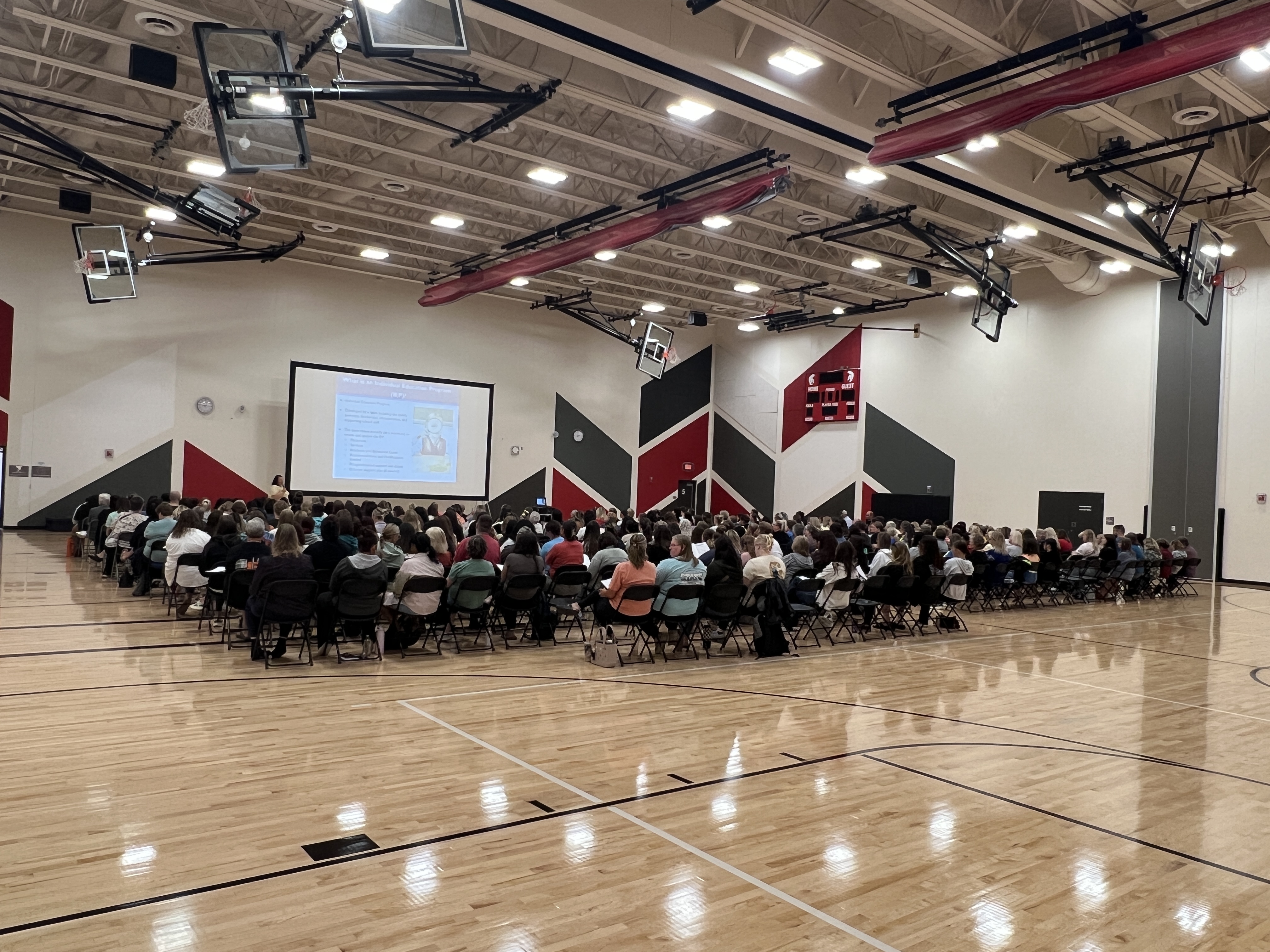 A large audience sits in chairs on a polished wooden gymnasium floor facing a projector screen, with a scoreboard and decorative wall panels visible in the background.