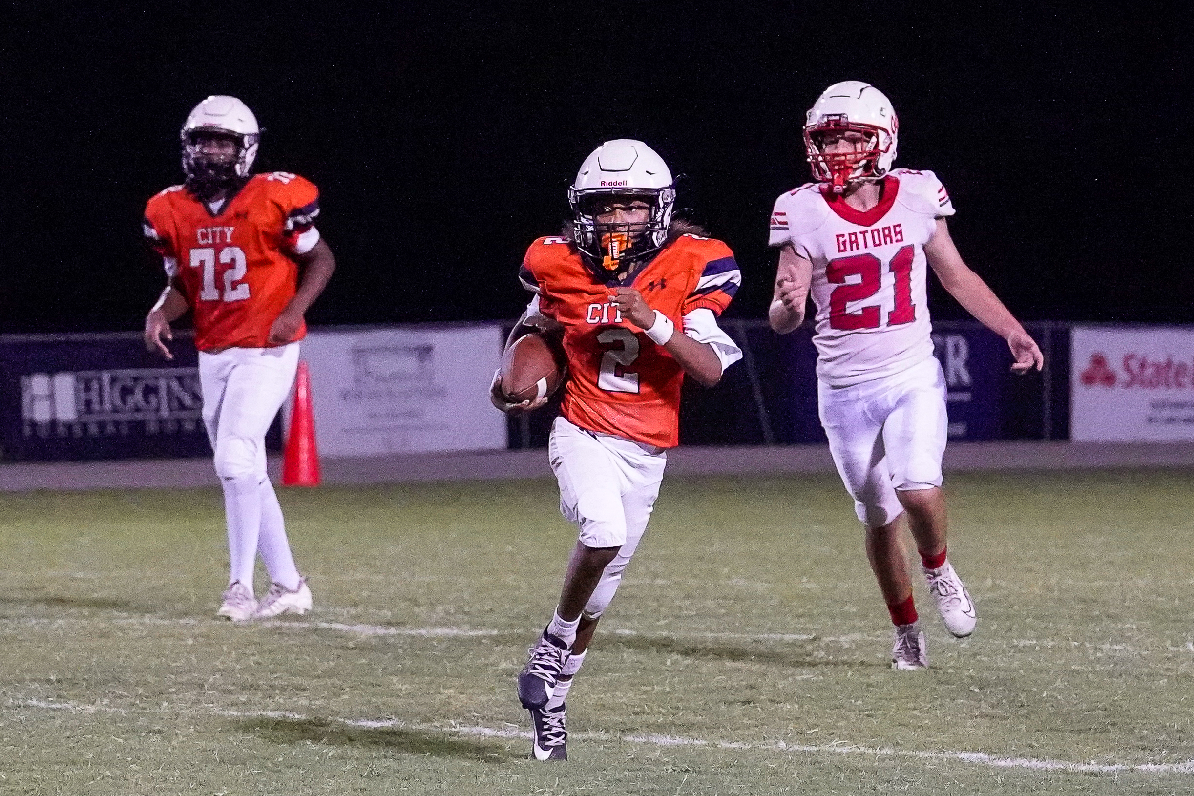 A young football player in an orange jersey runs with the ball on a field, while two other players in orange and white jerseys trail behind him, with a blurred background of stadium lights and advertisements.