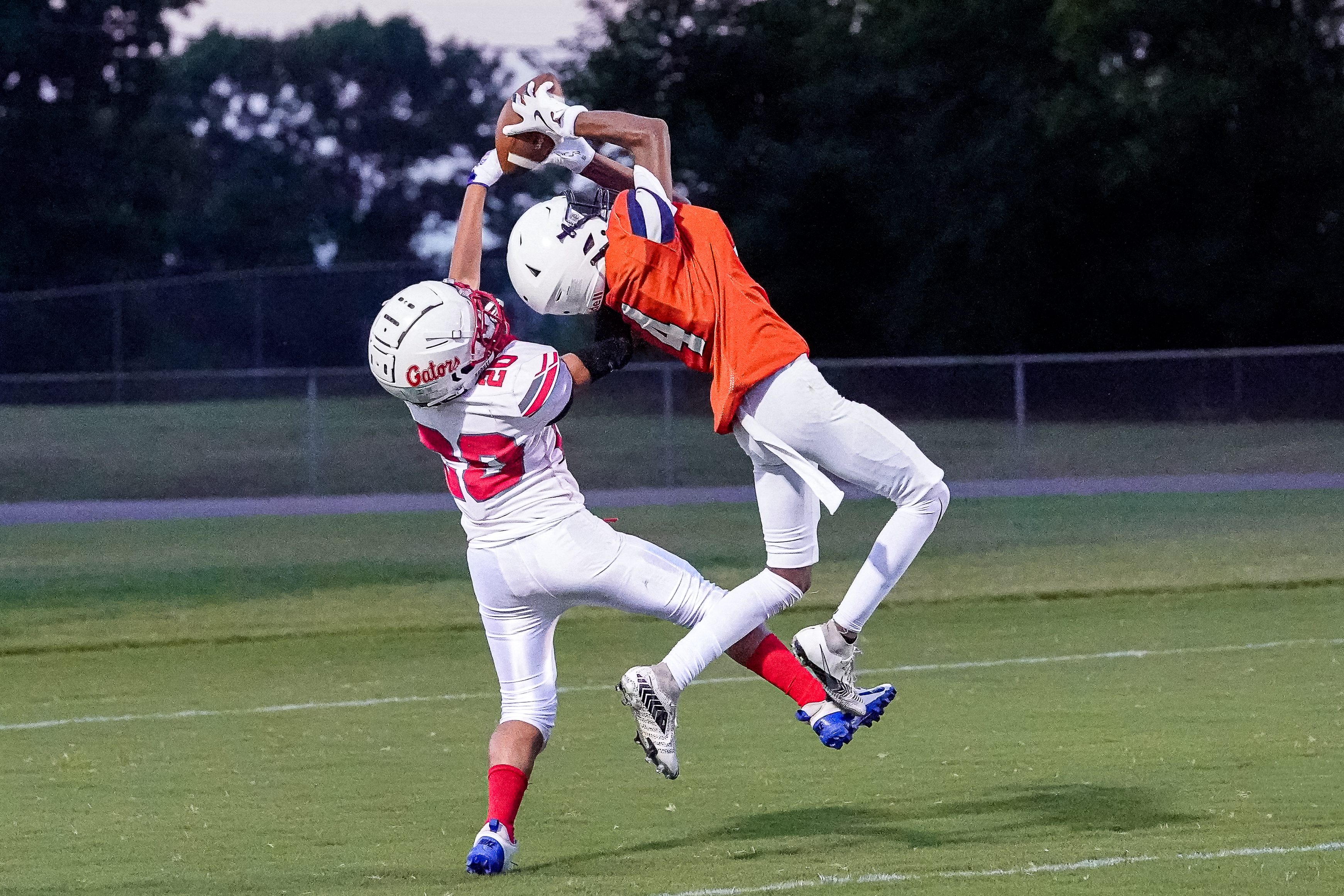 Two football players in mid-air reach for the ball against a backdrop of a grassy field and trees.