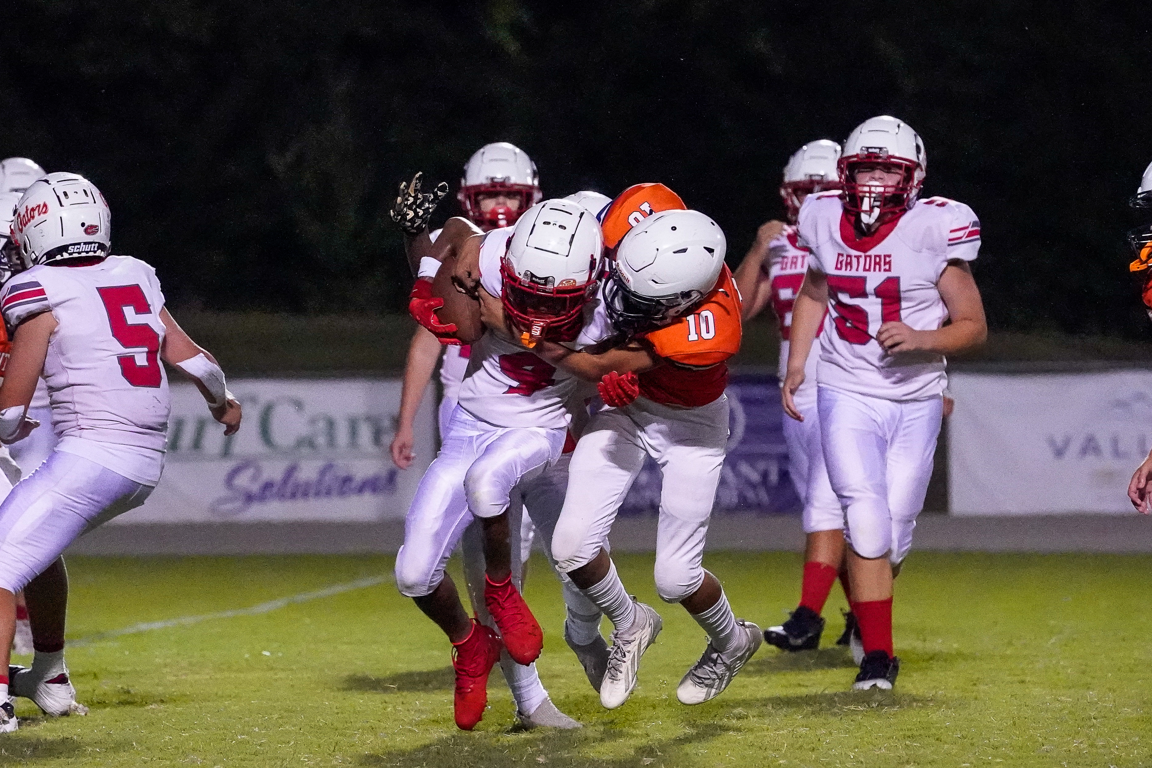Two football players in white and orange uniforms are tackling each other on a grassy field at night, with other players and stadium lights visible in the background.
