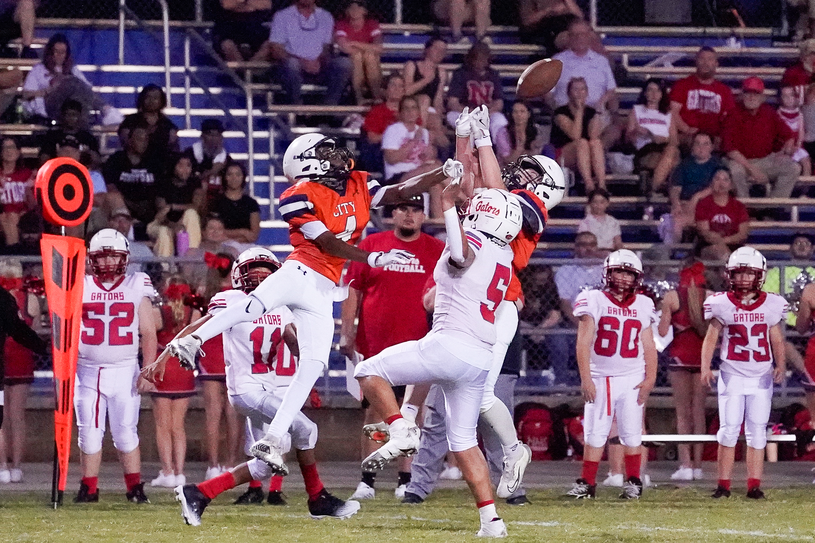 In the foreground, football players leap for a pass, while the background shows spectators in the stands.