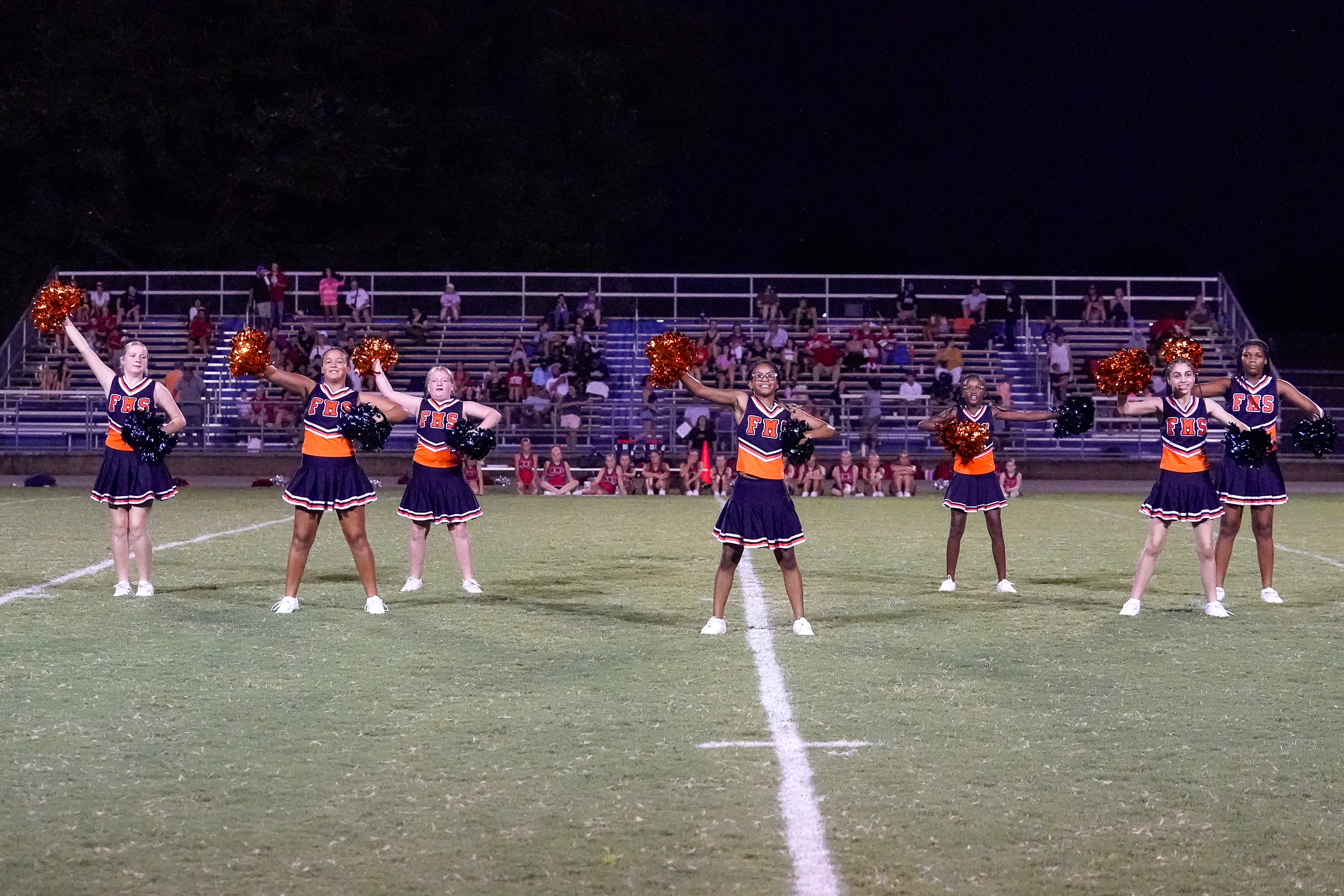 A group of cheerleaders in orange and black uniforms perform on a grassy football field in front of bleachers filled with spectators under a dark night sky.