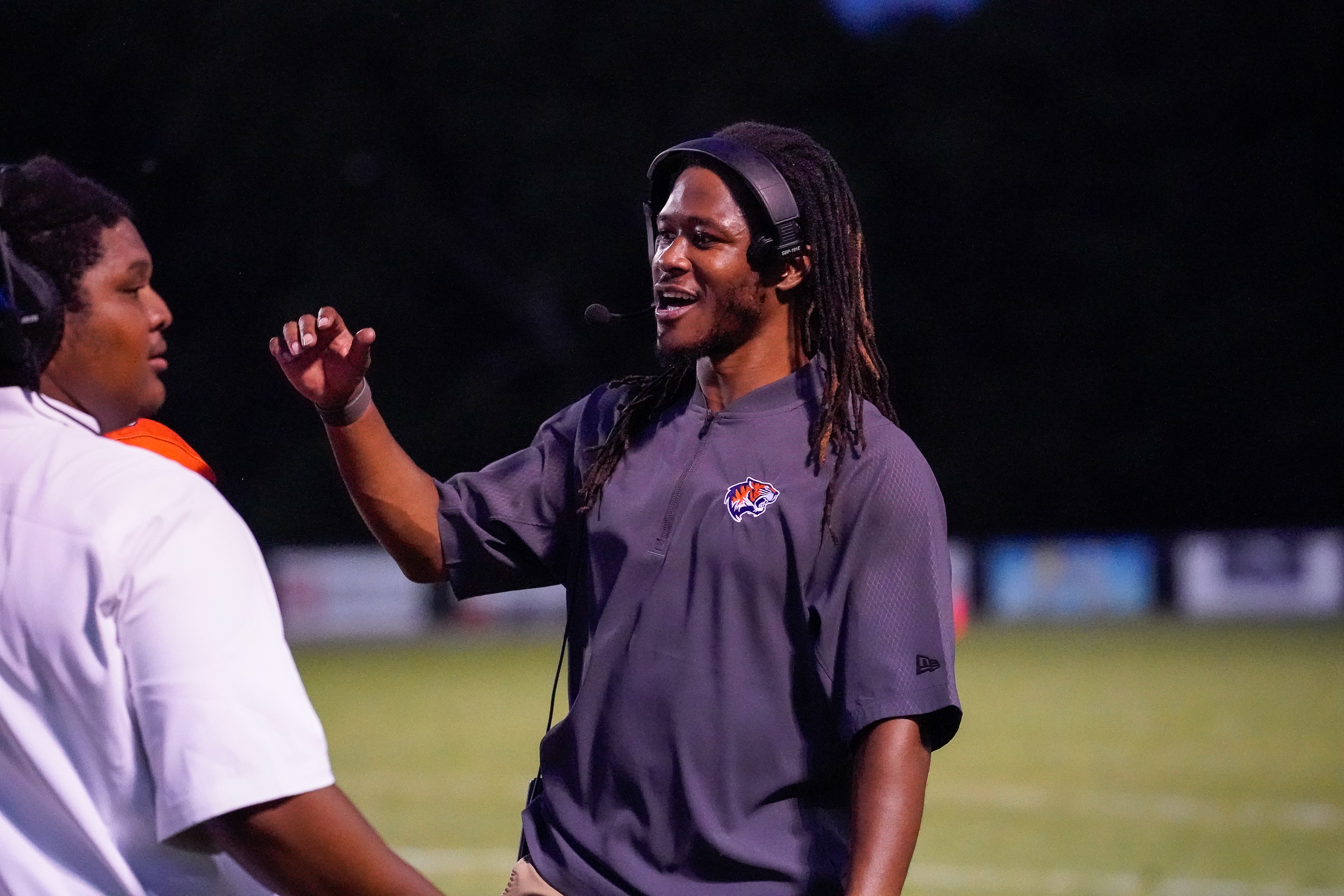 A coach with dreadlocks and headphones gestures while speaking to a player in the foreground, with a blurred field and lights in the background.