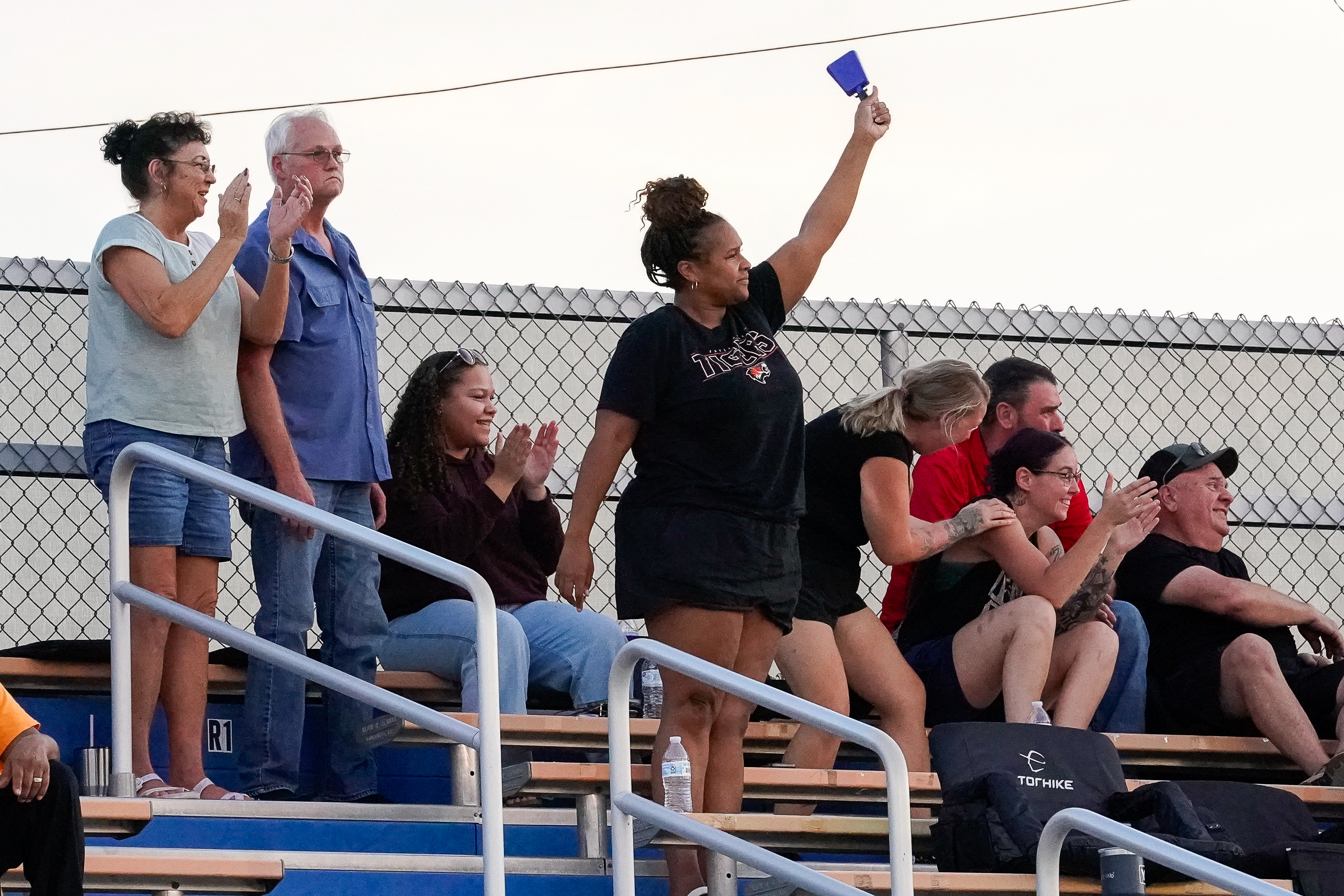 Spectators cheer from bleachers in front of a chain-link fence under a pale sky.