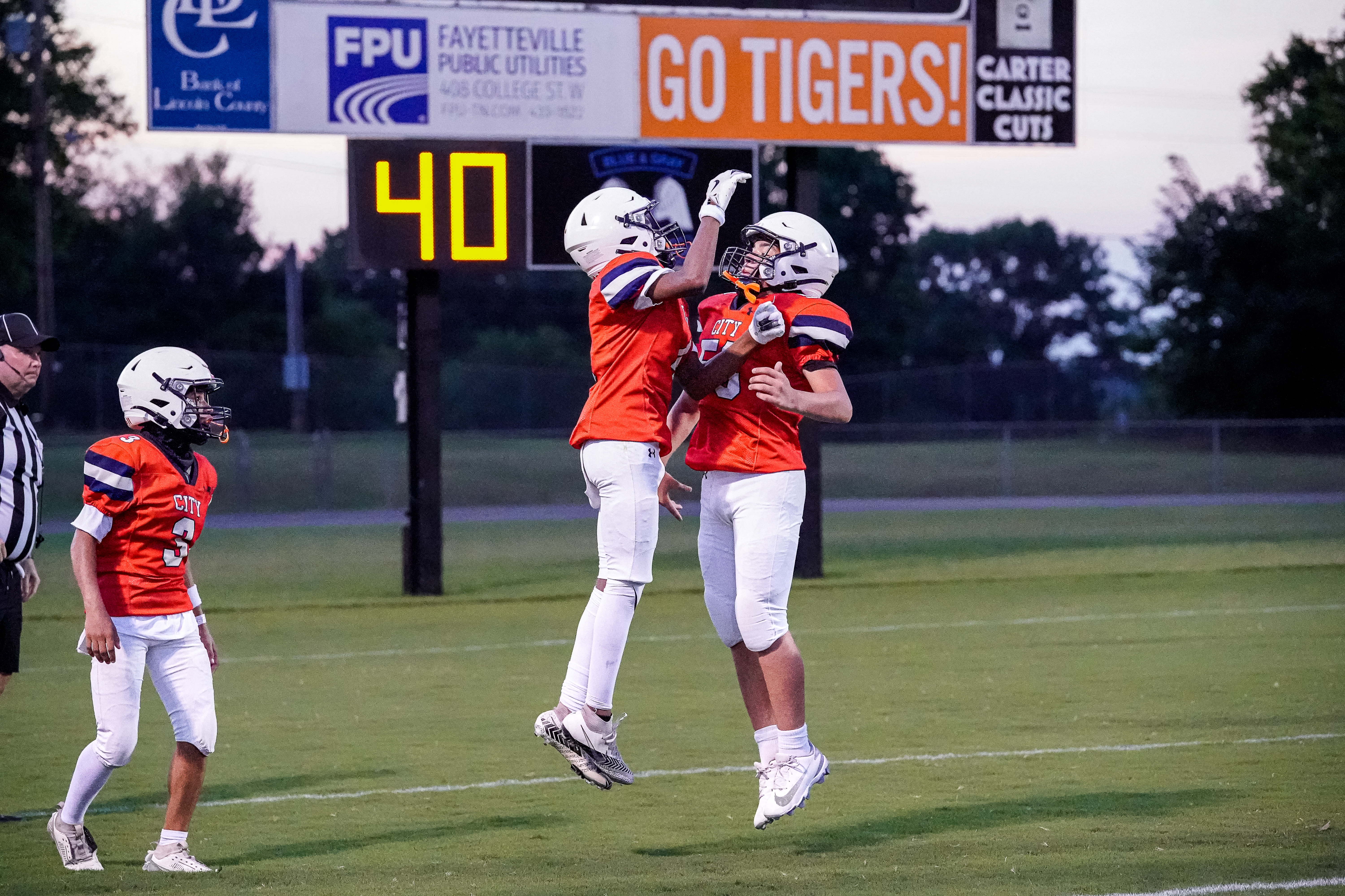 Two young football players in orange and white uniforms jump in celebration on a green field, with a scoreboard displaying "40" and "GO TIGERS!" in the background.
