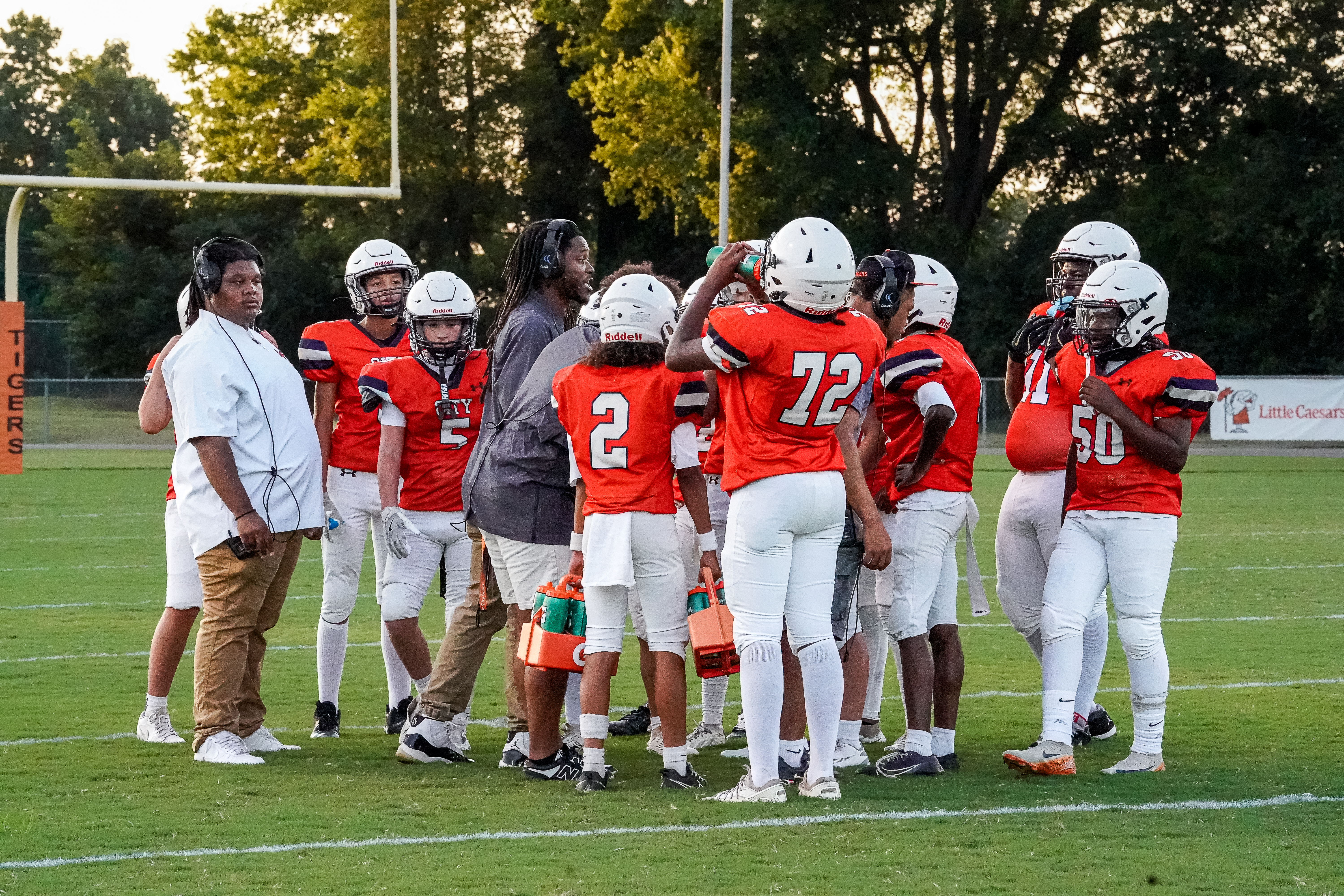 A group of young football players in orange jerseys huddle on a green field with goalposts in the background.