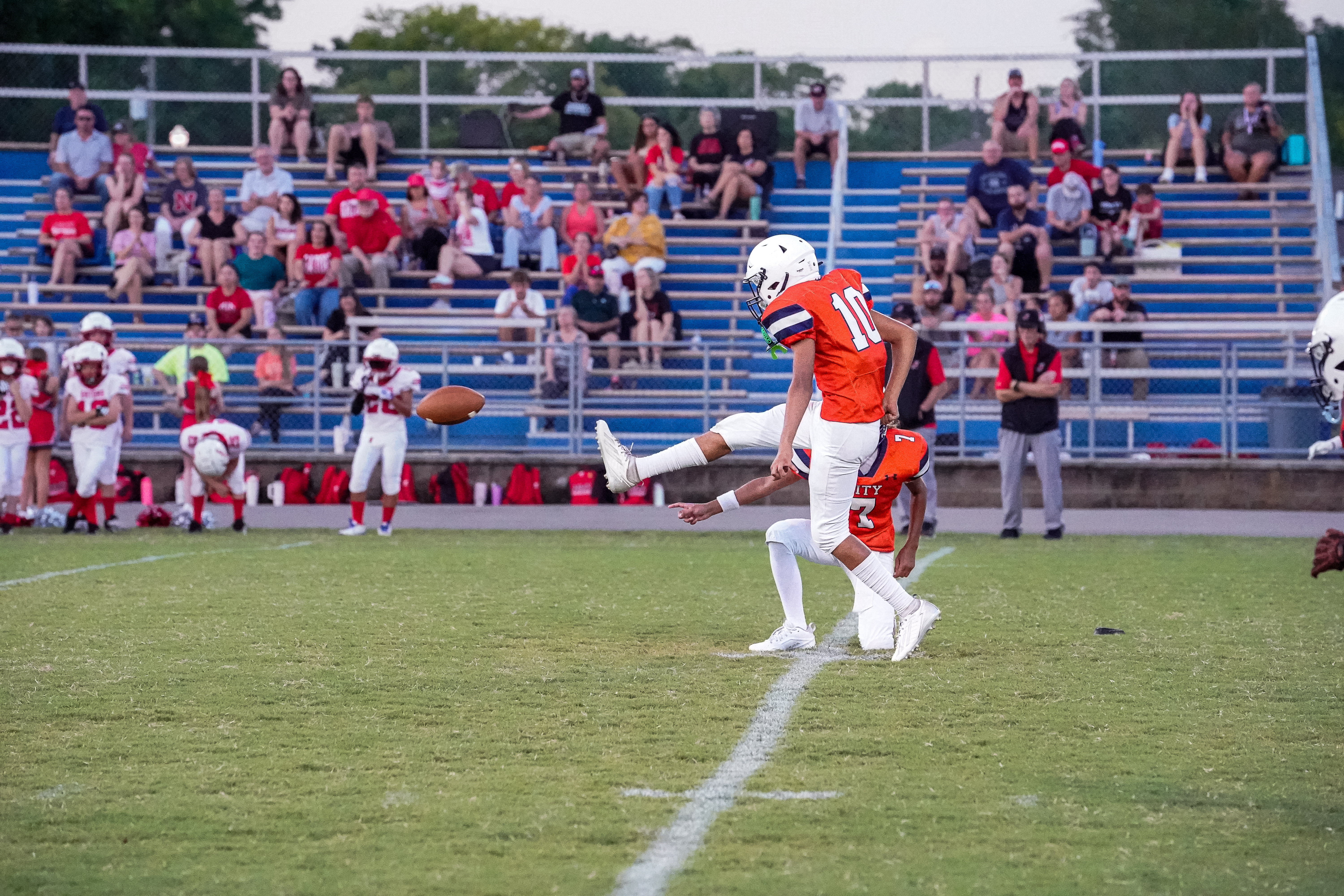A football player in an orange jersey kicks the ball on a grassy field with spectators in bleachers in the background.