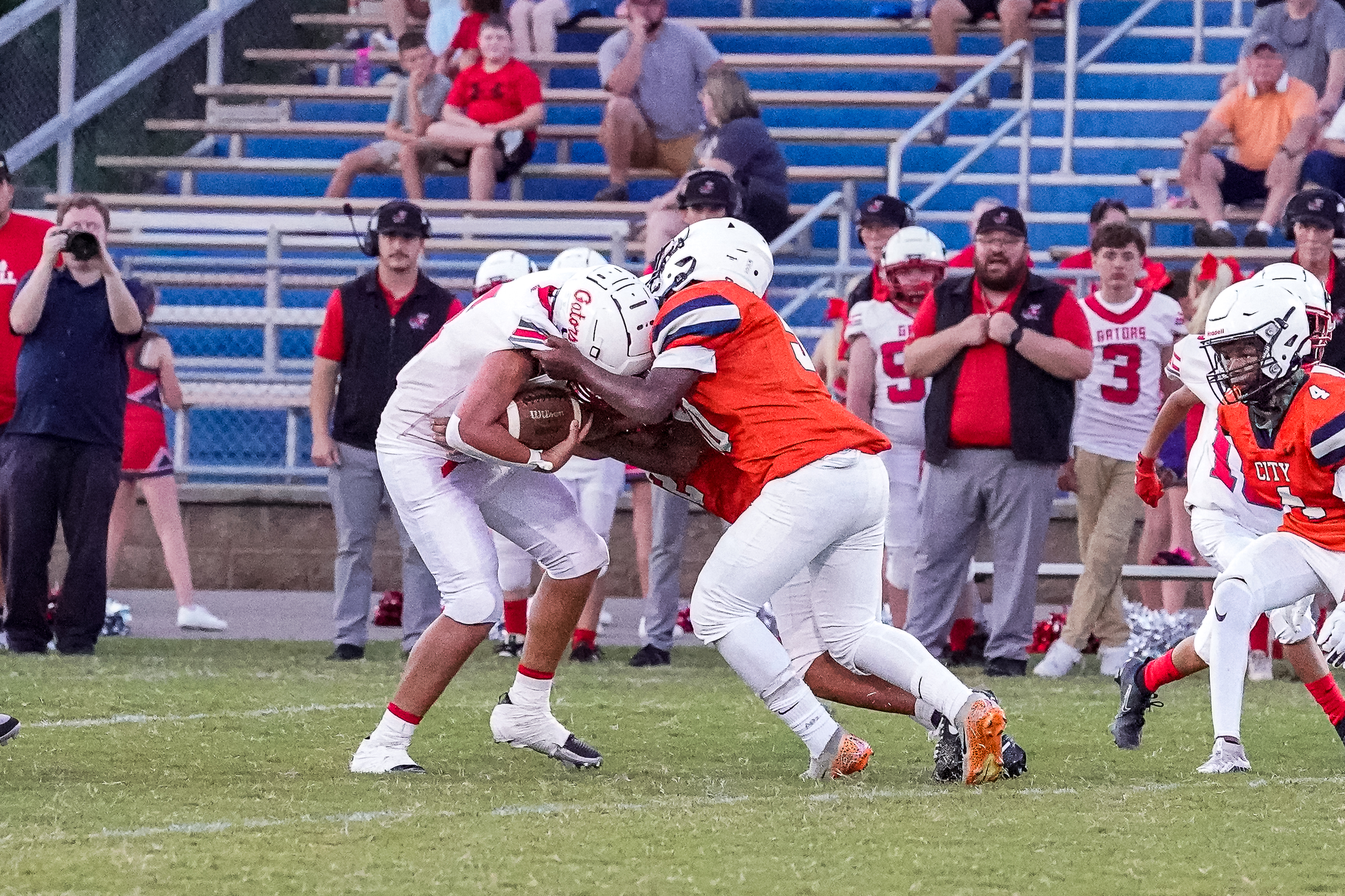 Two football players in white and orange uniforms tackle a player with a football in the foreground, while spectators watch from bleachers in the background.