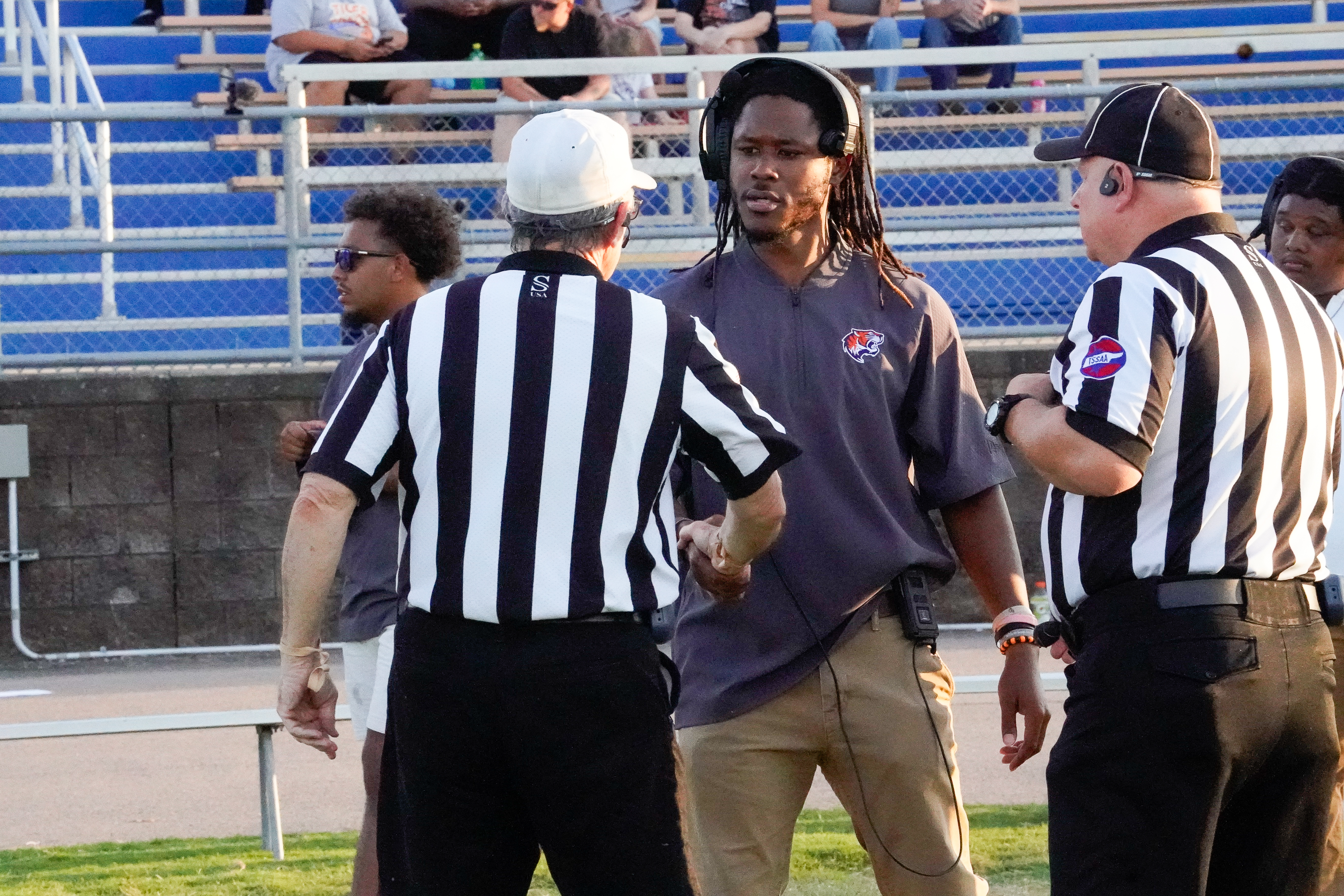 Three men in referee uniforms and one man in a grey shirt with headphones are on a grassy field in front of bleachers filled with spectators.