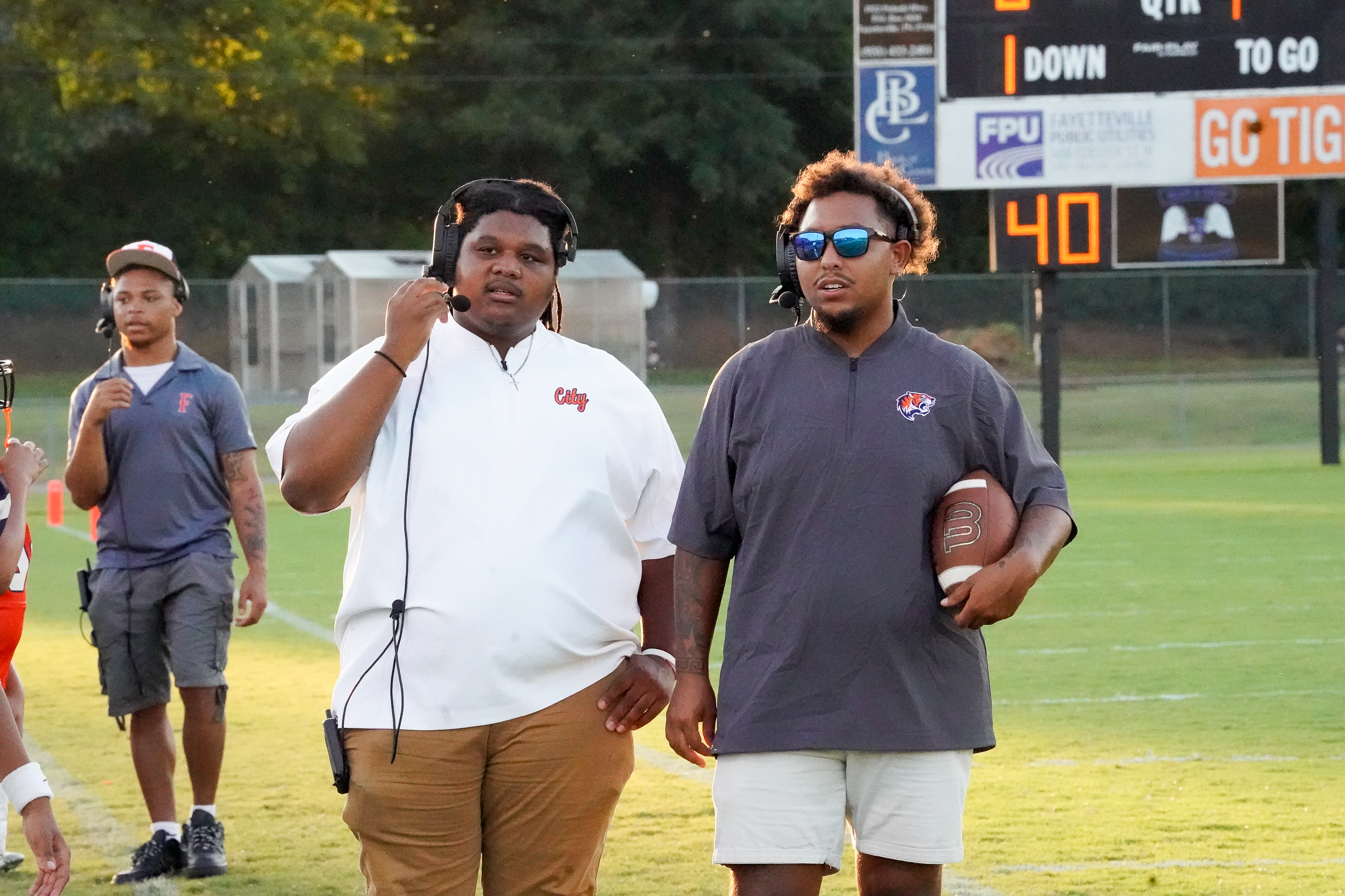 Two coaches stand on a football field in the foreground, while a scoreboard and trees are visible in the background.