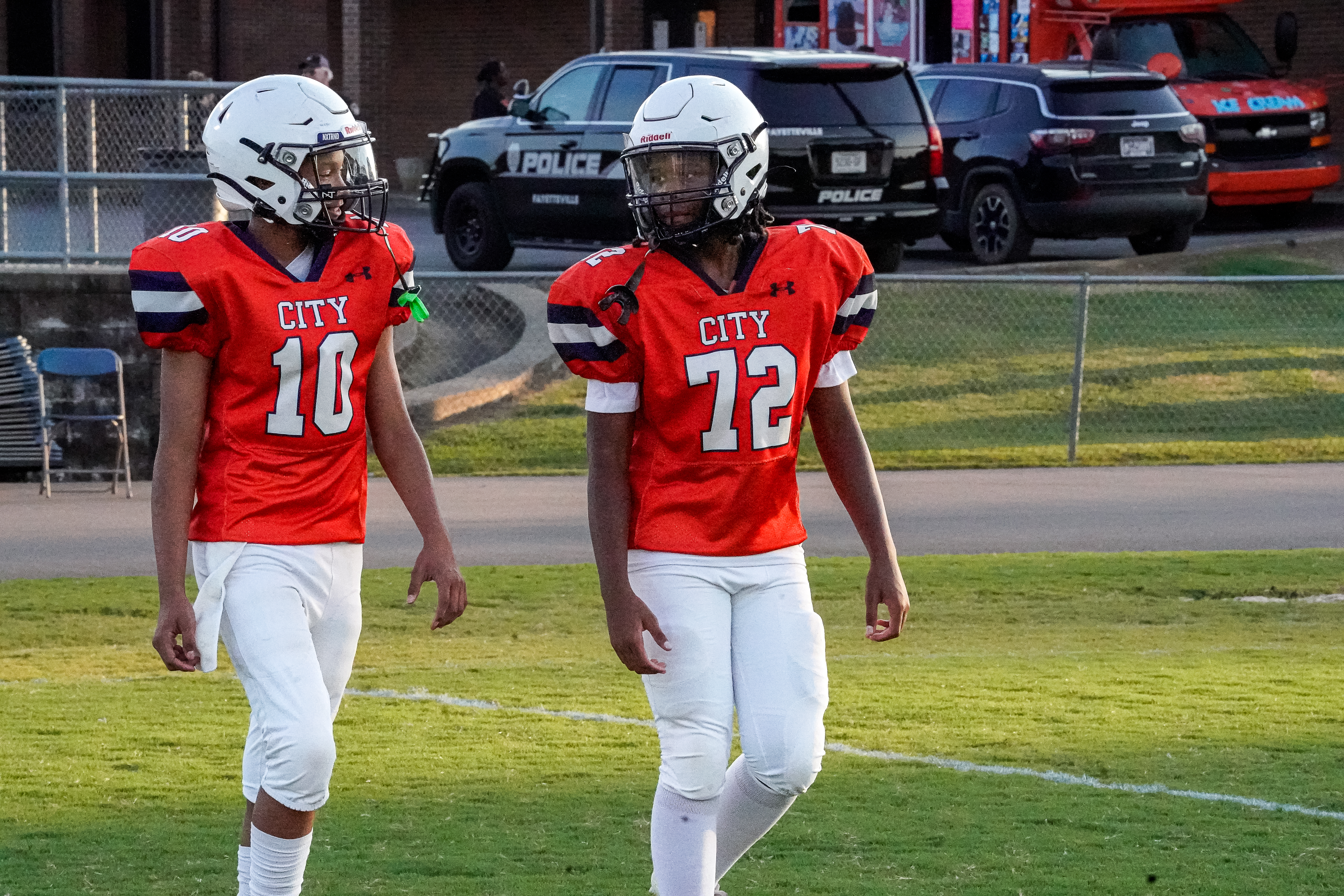 Two young football players in orange jerseys walk across a grassy field with police vehicles in the background.