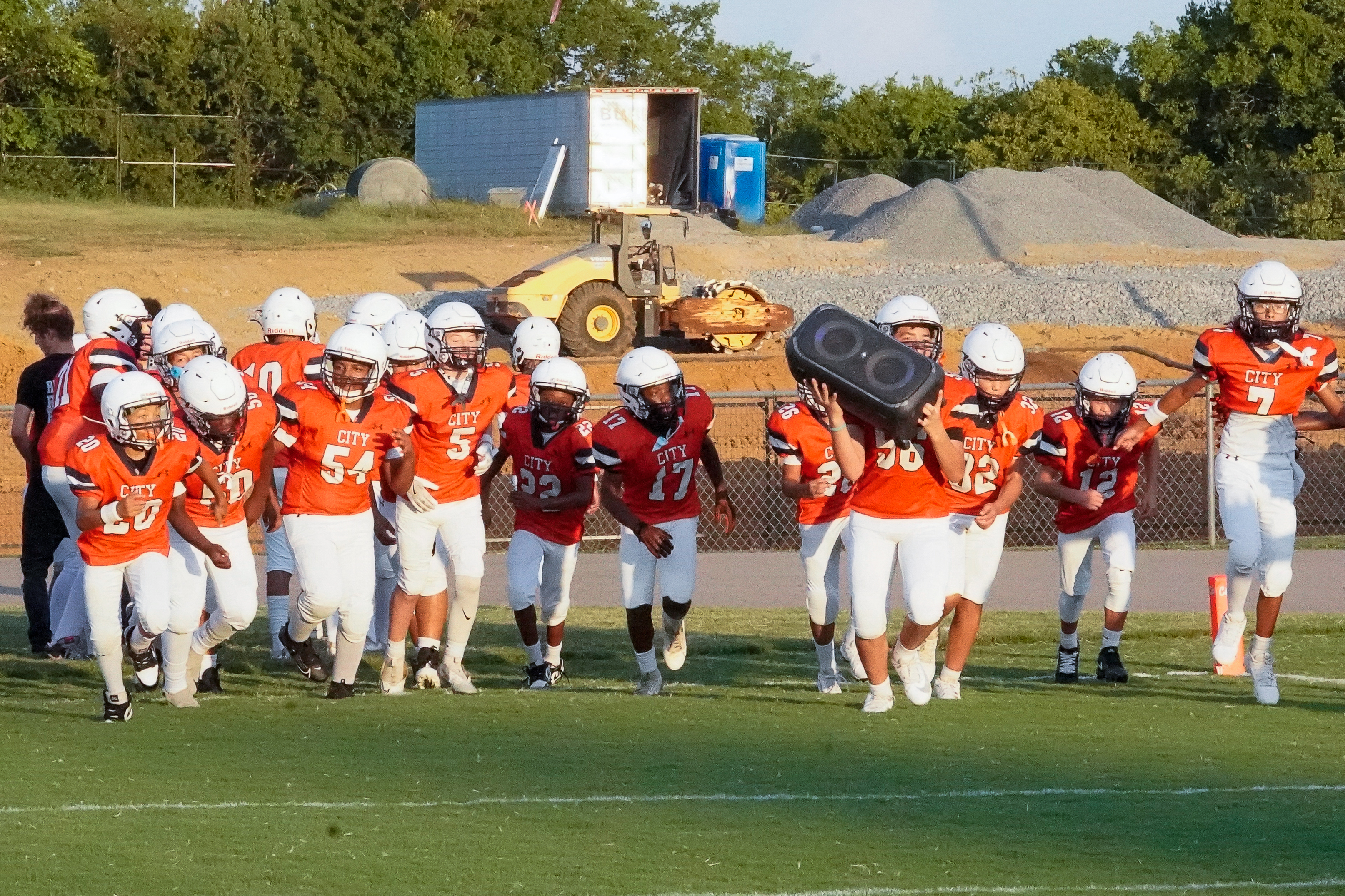A group of young football players in orange and white uniforms run onto a field in the foreground, while a construction site with heavy machinery and piles of dirt is visible in the background.