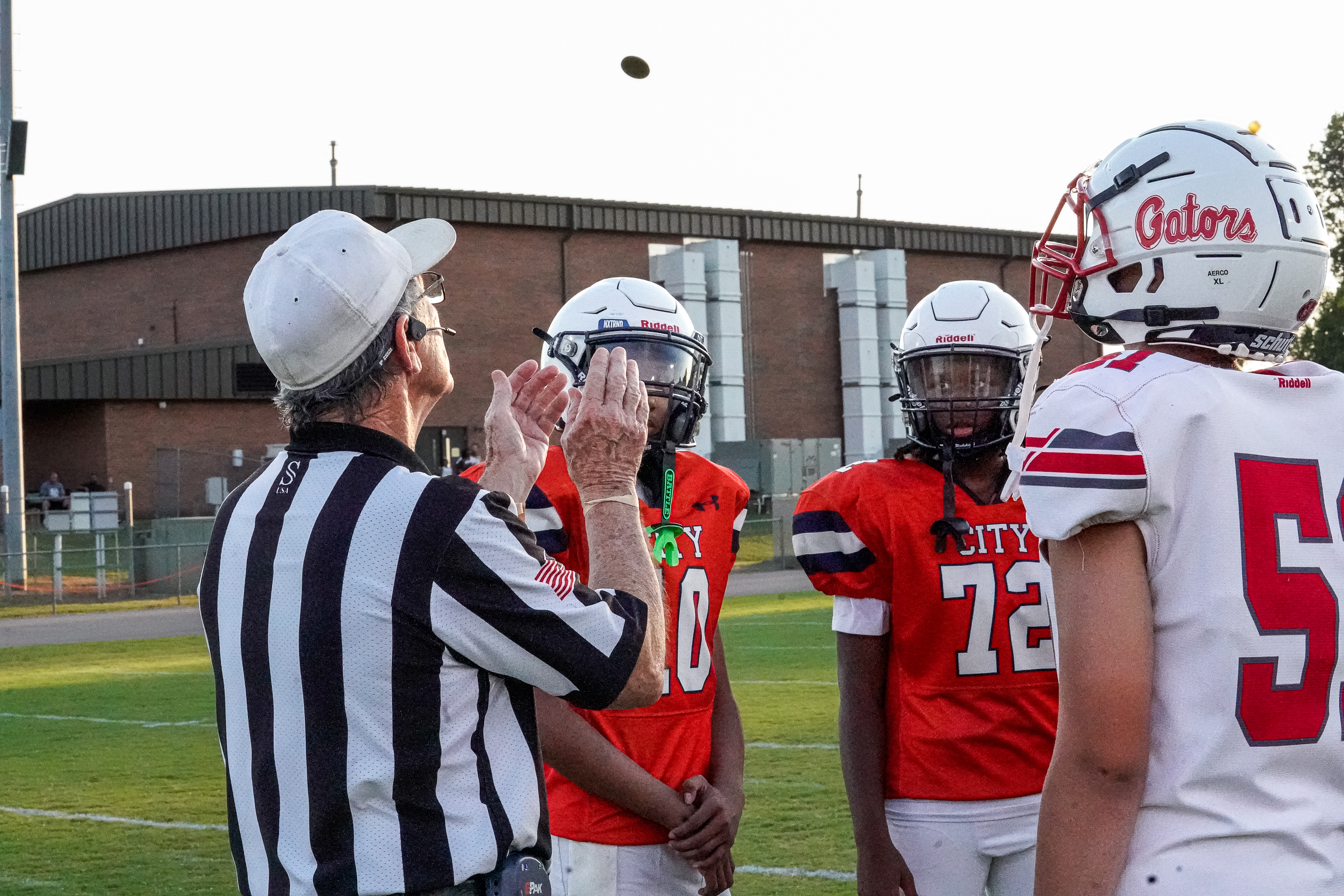 A referee in a striped shirt tosses a football in the air as two young football players in orange jerseys and helmets stand ready on a field, with a building in the background.