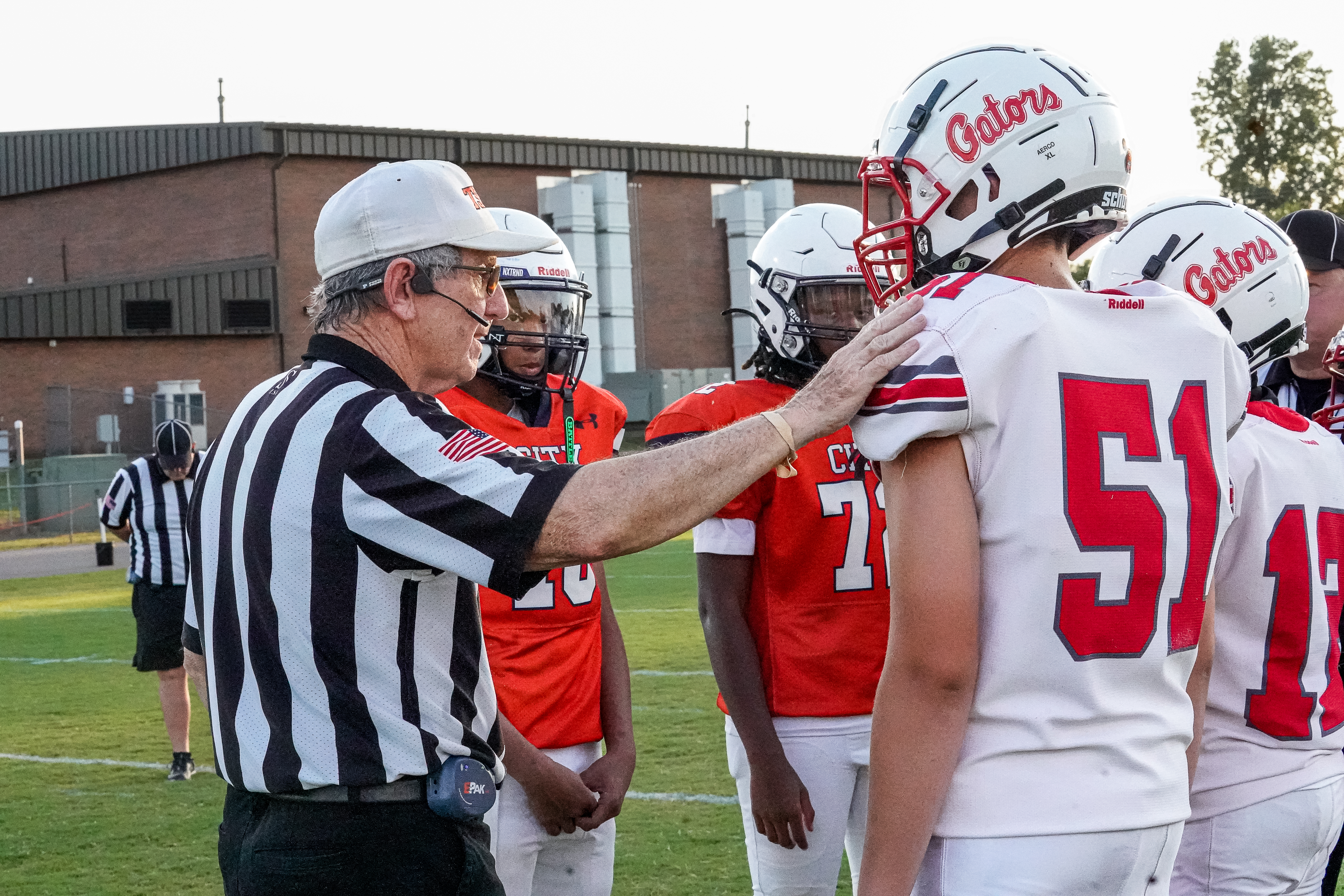 A referee in a striped shirt talks to football players in orange and white jerseys on a grassy field with a building in the background.