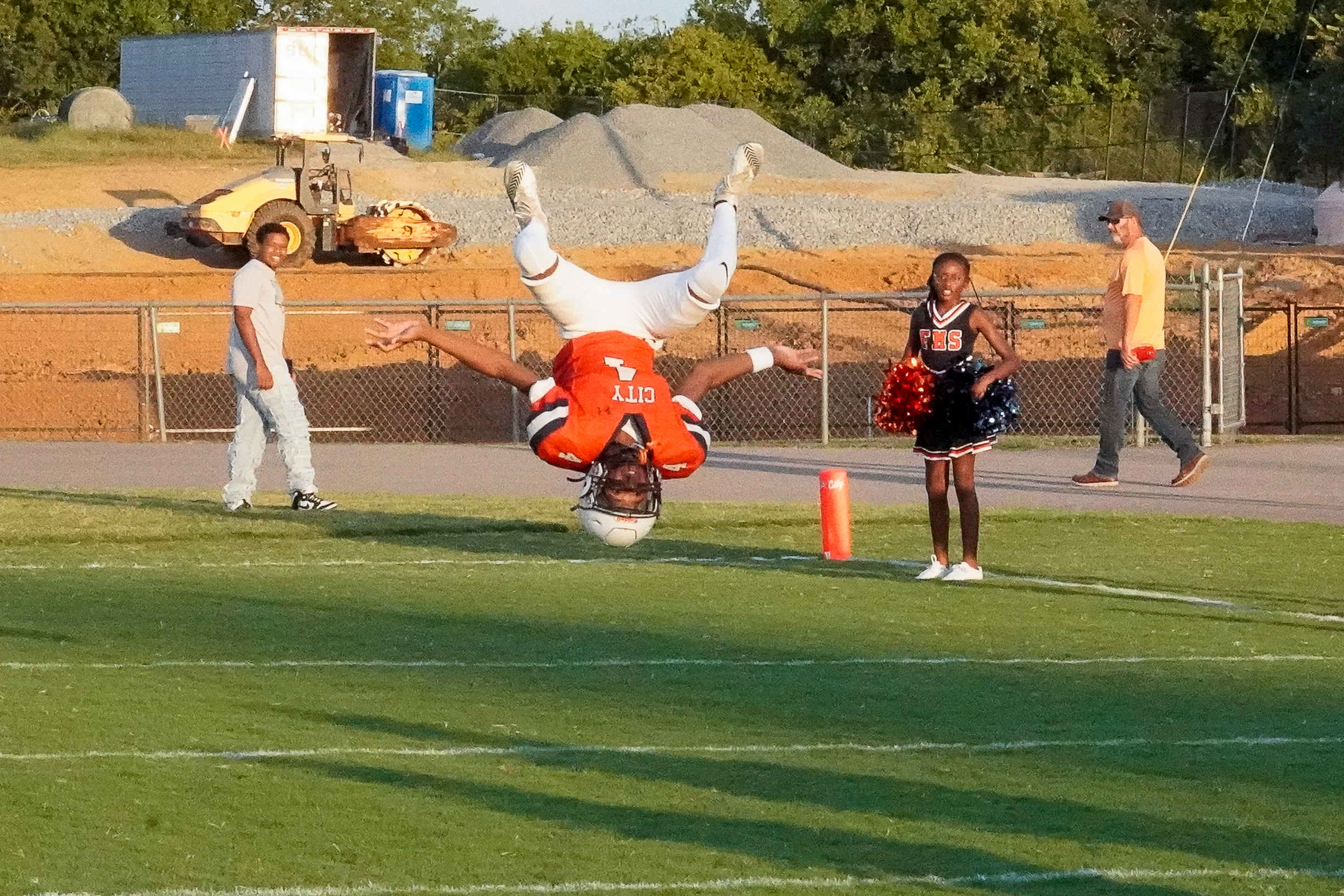 A football player performs a backflip on a grassy field in the foreground, while construction equipment and onlookers are visible in the background.