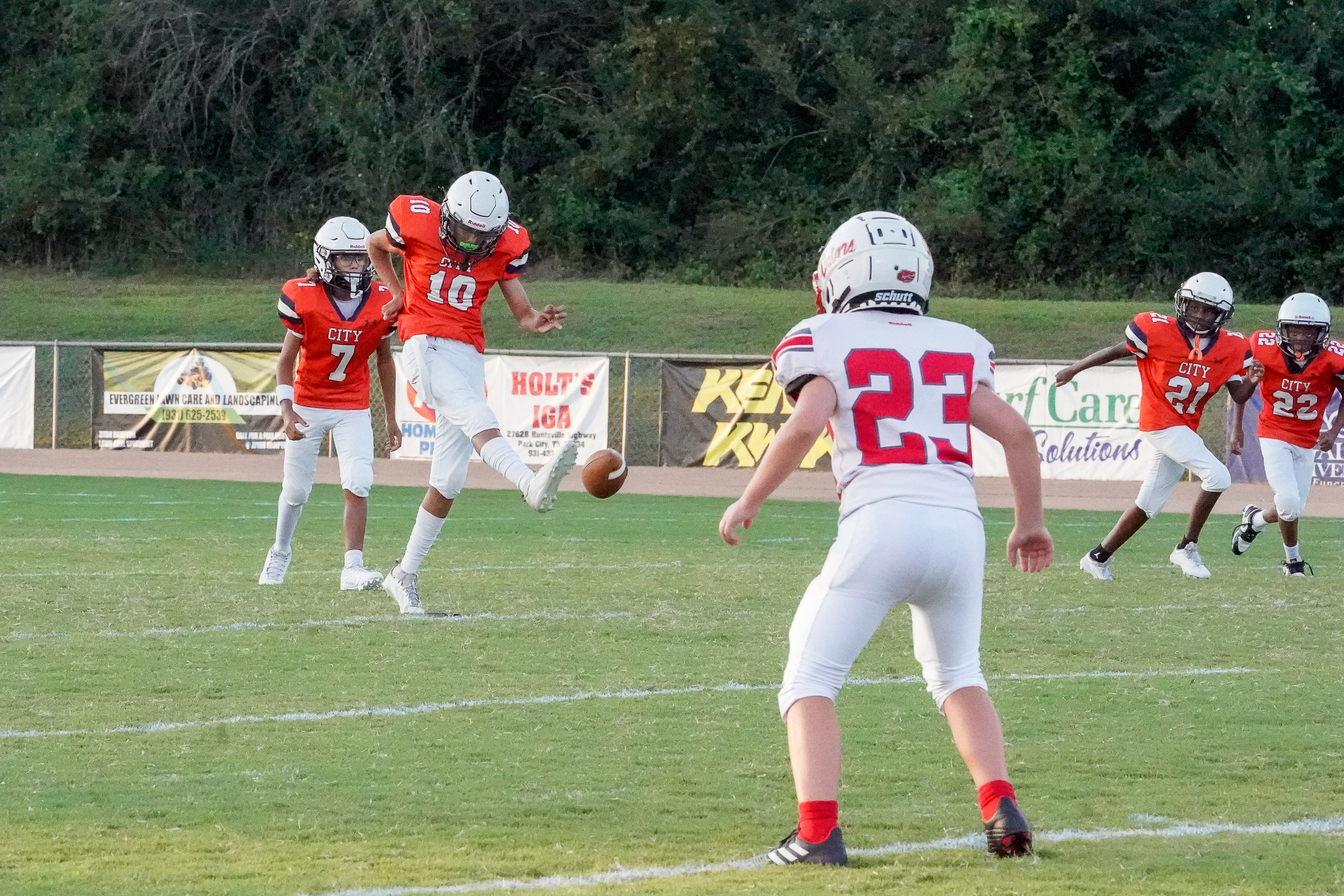 A football player in an orange uniform kicks the ball on a green field in the foreground, with other players and a stadium in the background.