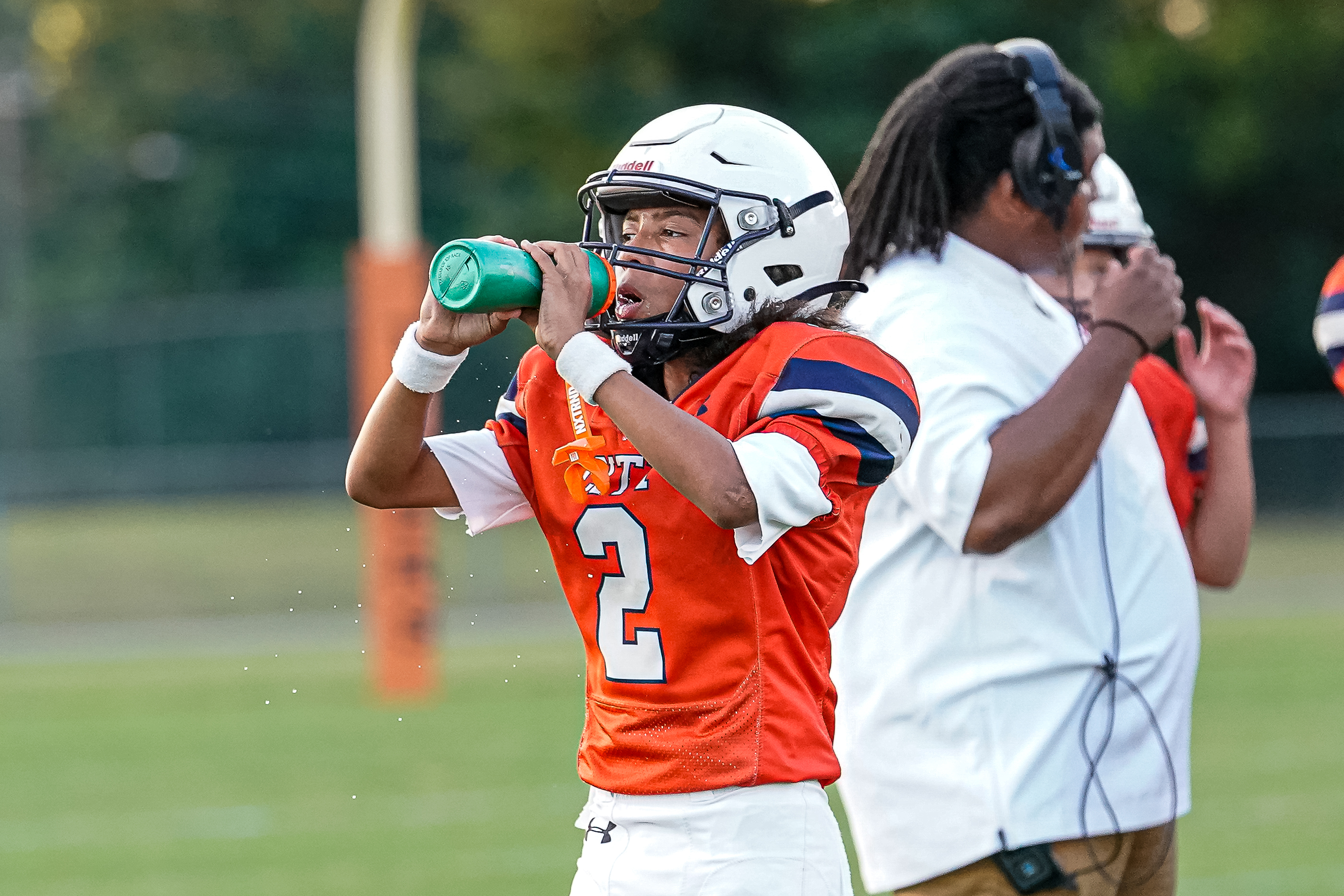 A young football player in an orange jersey drinks from a green water bottle in the foreground, while a coach with headphones stands beside him against a blurred background of a field and goalpost.