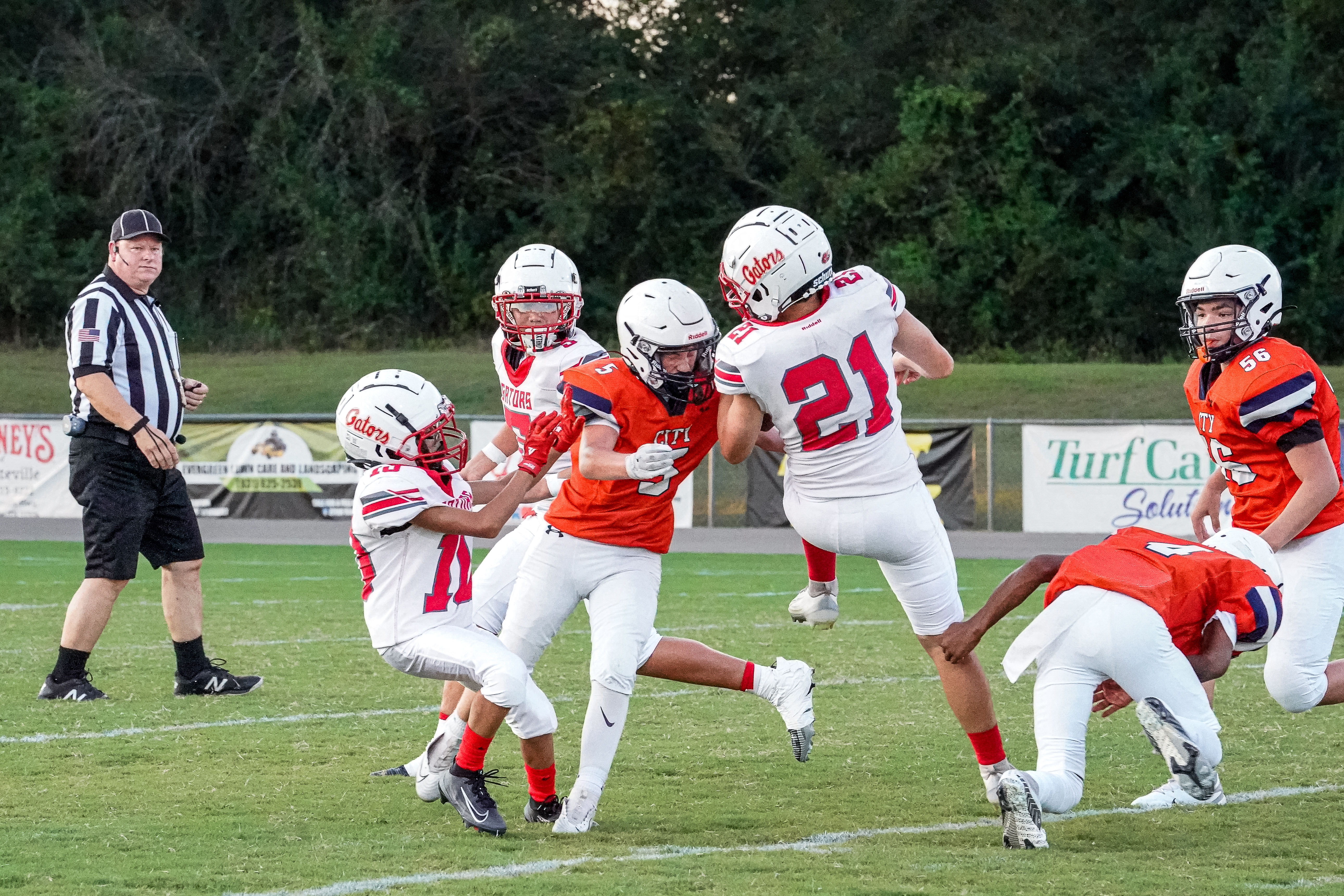 A football player in an orange jersey is tackled by opponents in white jerseys on a green field, with a referee watching in the background.