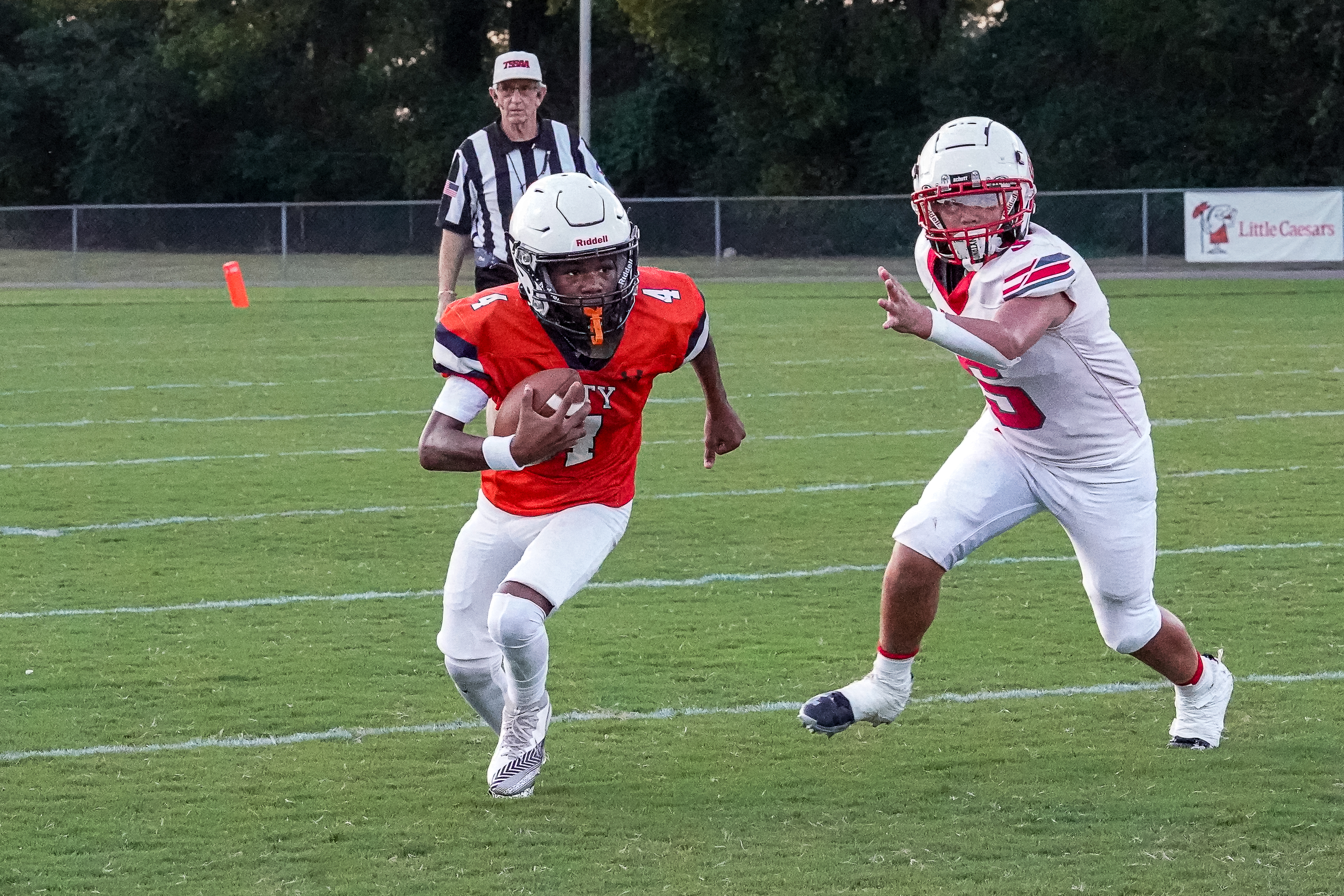 A football player in an orange jersey runs with the ball on a green field, pursued by a player in a white jersey, with a referee and a fence in the background.