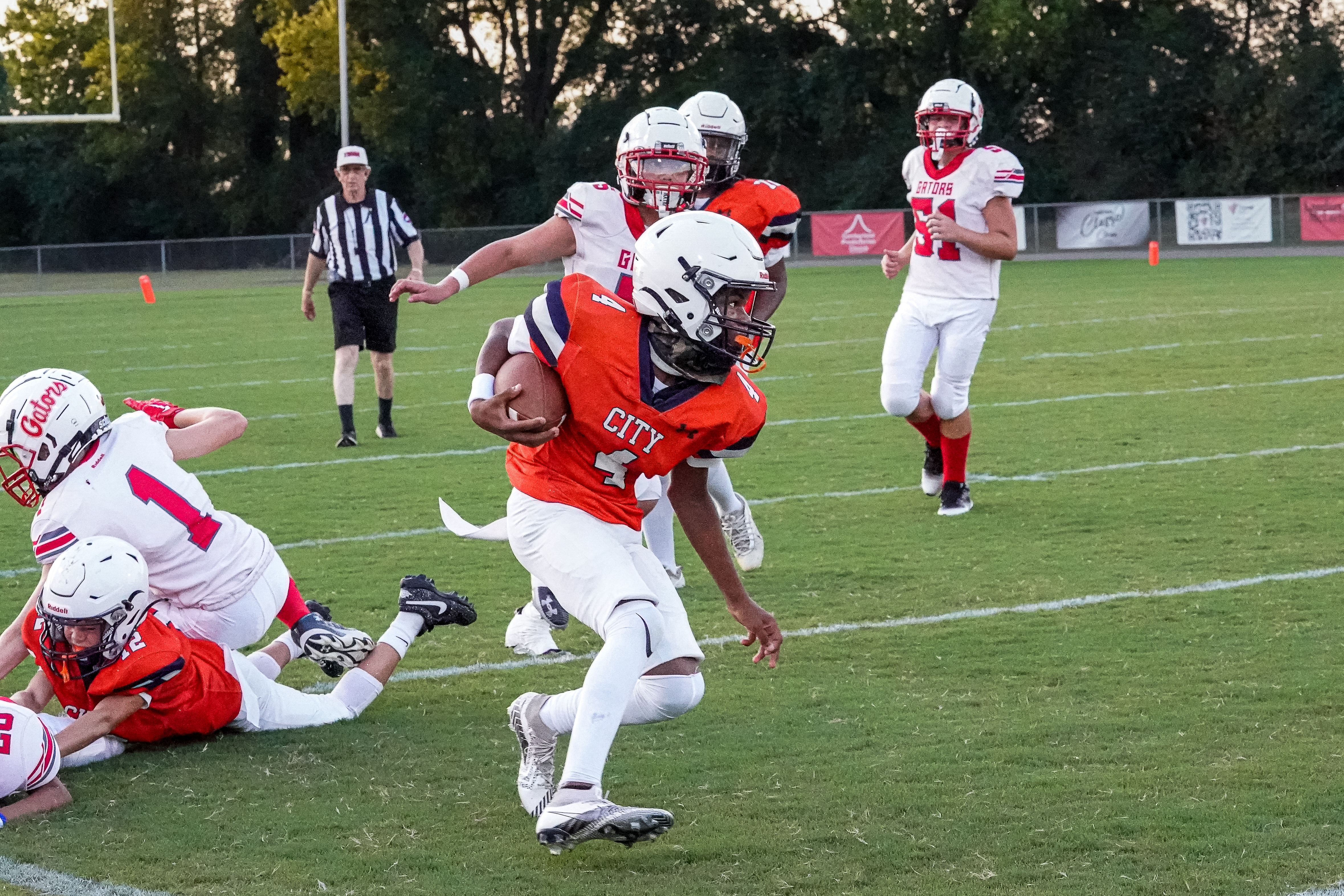 An orange-clad football player runs with the ball past defenders on a grassy field with a referee and spectators in the background.