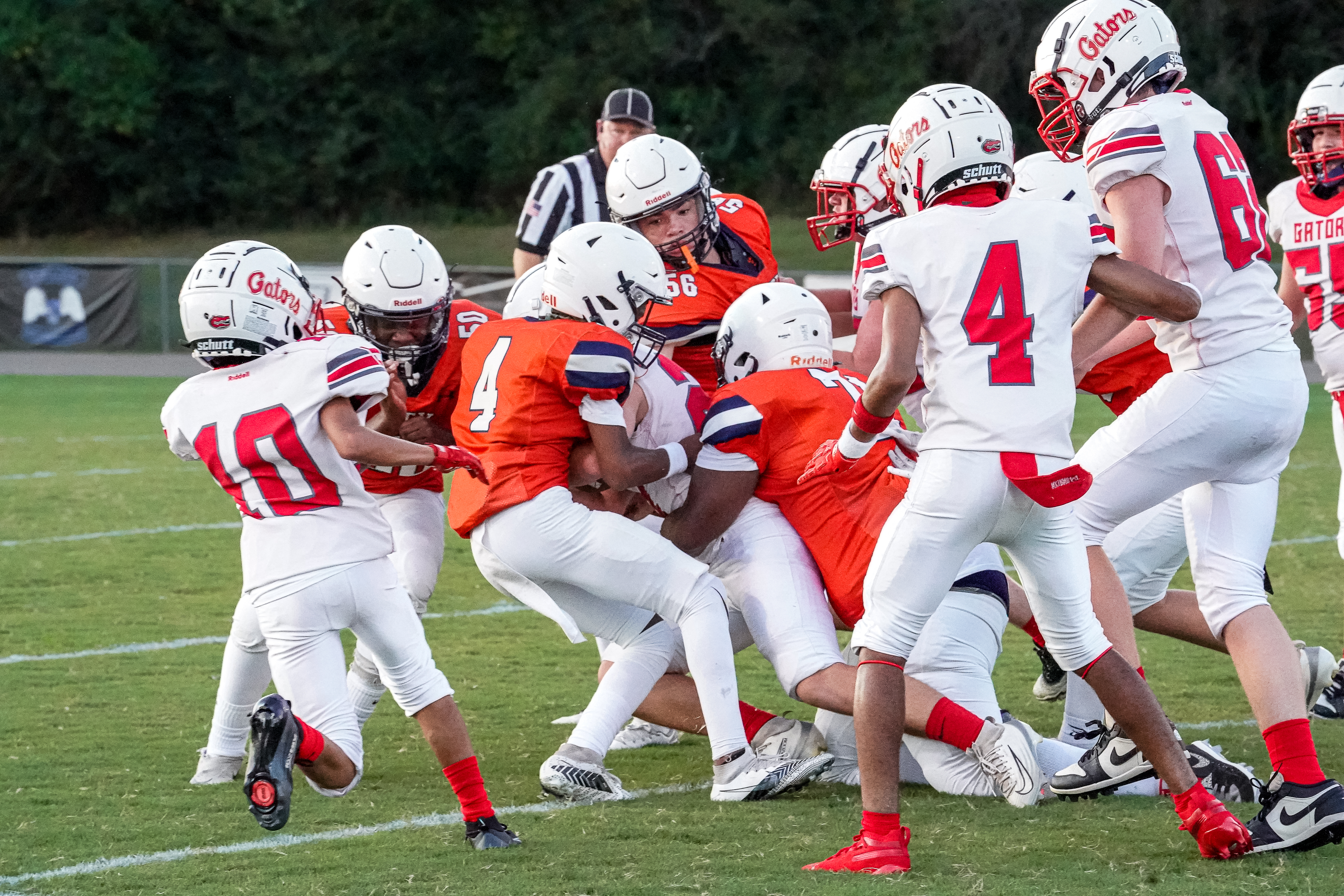 A group of young football players in white and orange uniforms are engaged in a play on a grassy field, with a referee in the background.