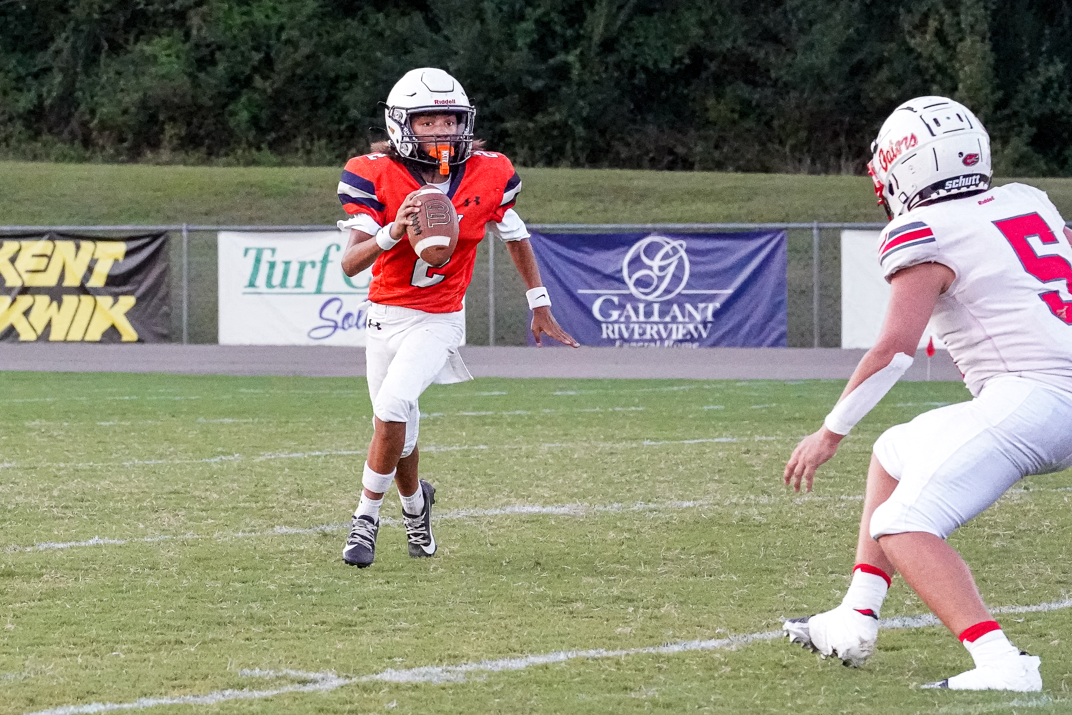 A young football player in an orange jersey runs with the ball on a grassy field, with banners and trees visible in the background.