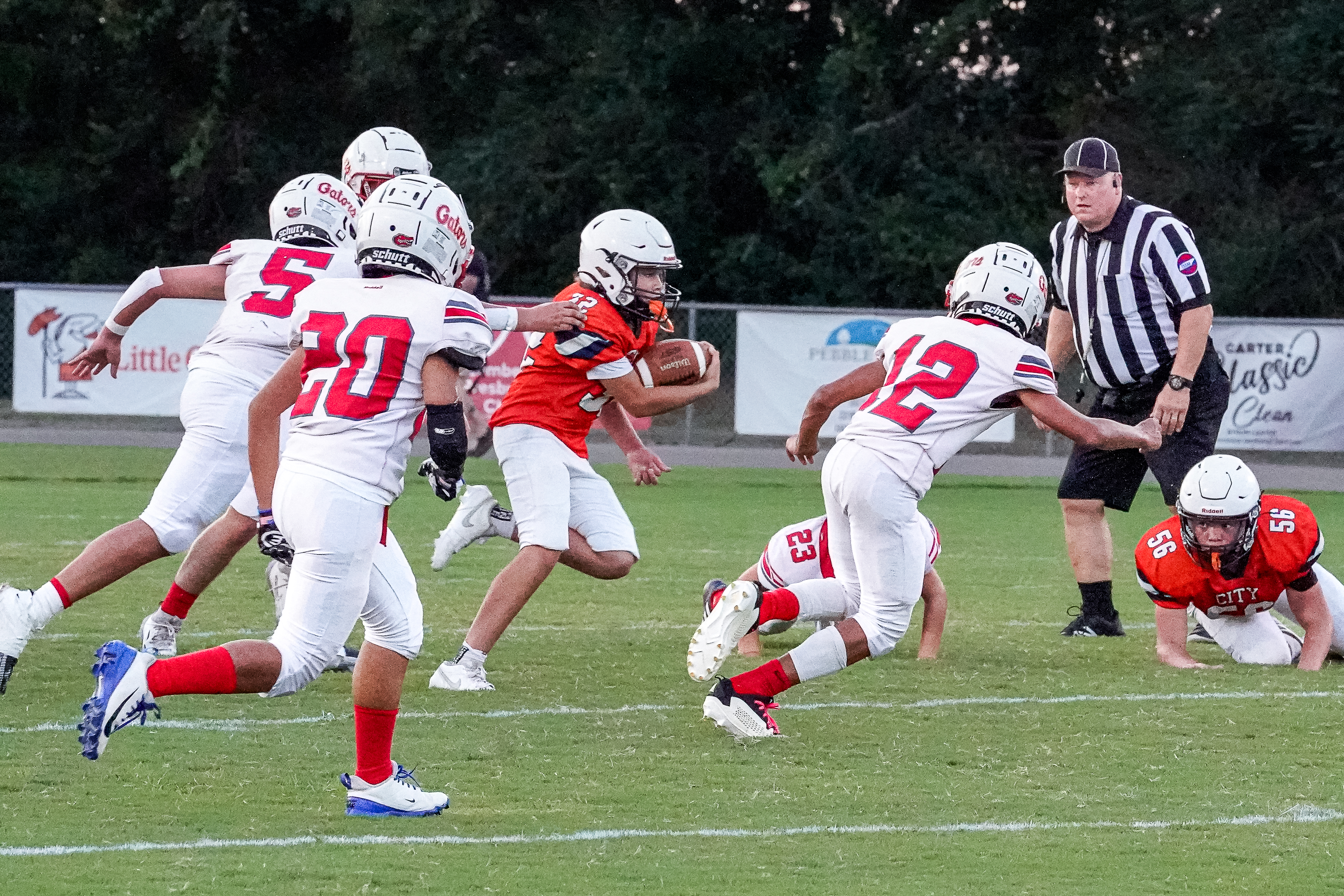 In the foreground, a young football player in an orange jersey runs with the ball while being pursued by players in white jerseys, and in the background, a referee watches the play unfold on a grassy field with trees and a fence.