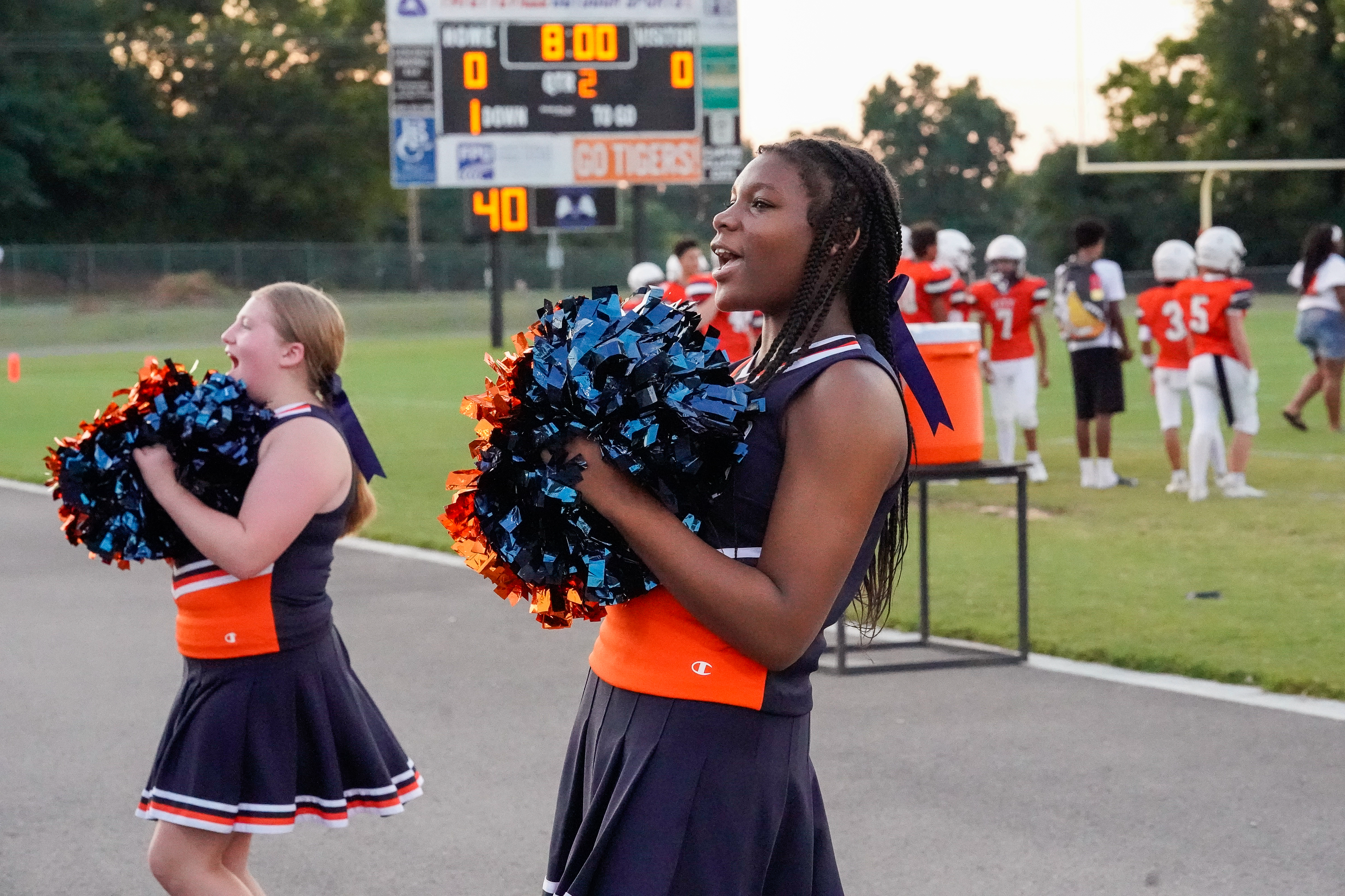 Two young cheerleaders in navy and orange uniforms with pom-poms in the foreground cheer at a football game, with a scoreboard and players in the background.