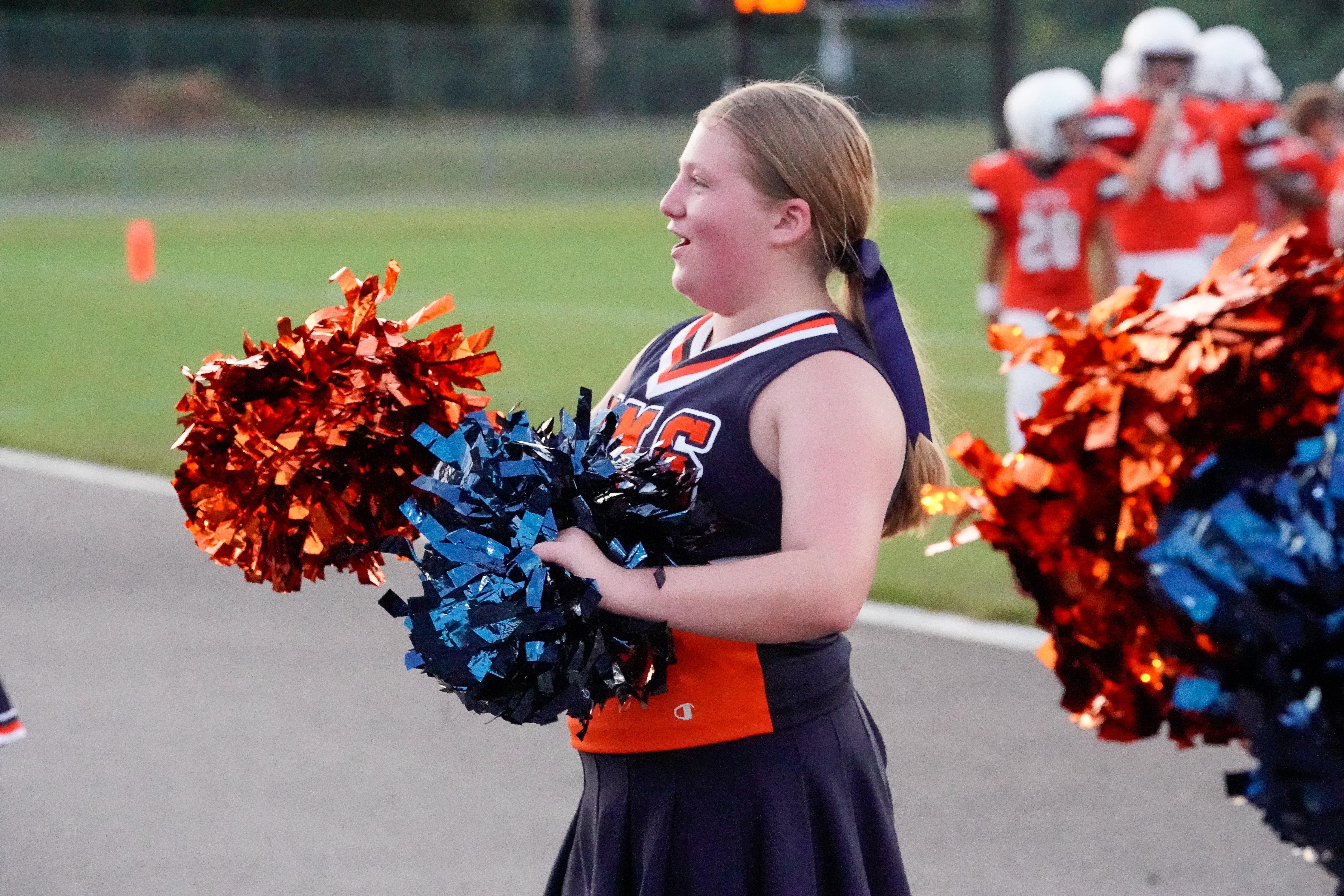 A young cheerleader with orange and blue pom-poms smiles as football players in orange jerseys stand in the background.