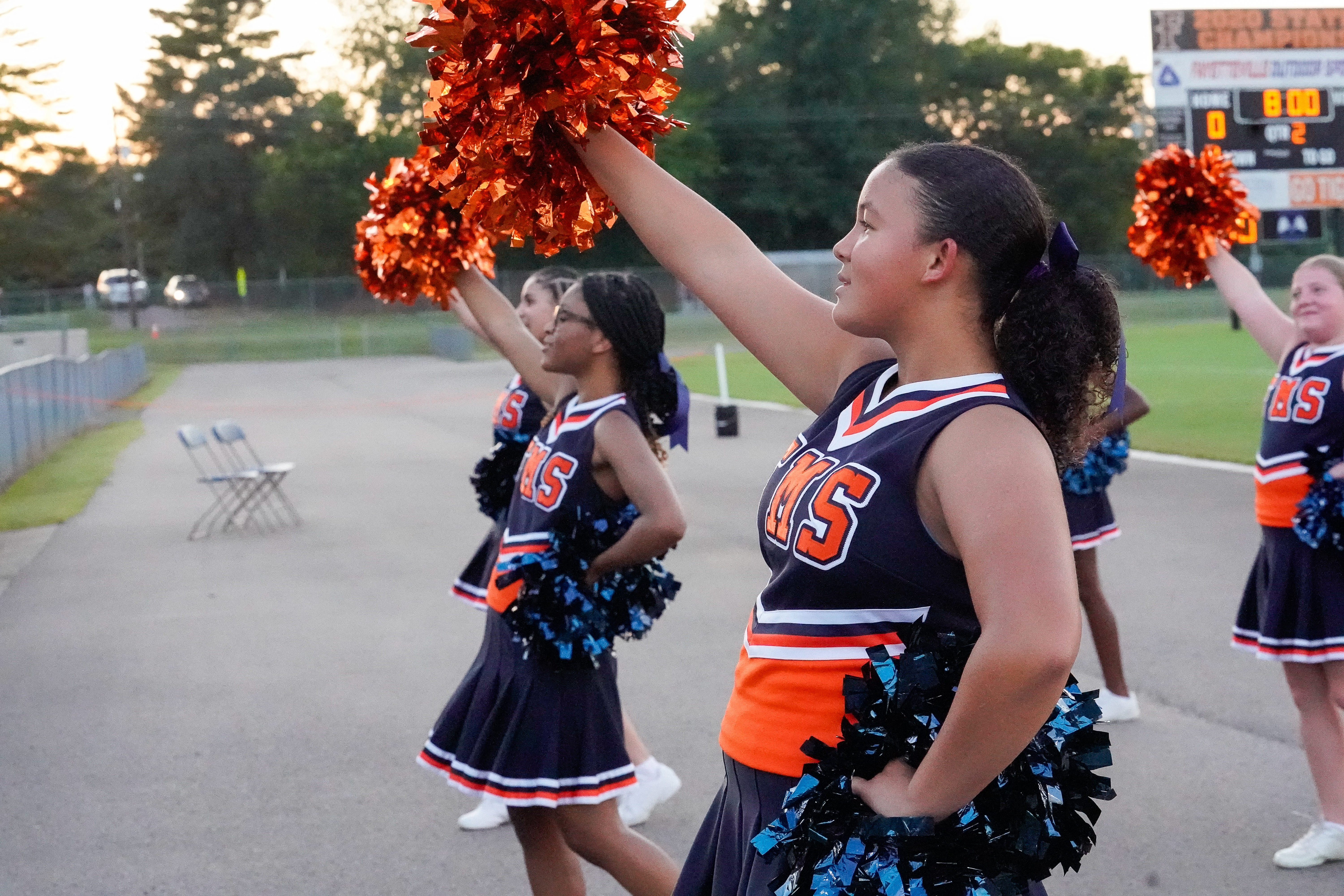 Cheerleaders in uniform with orange and blue pom-poms stand on a track in front of a football stadium scoreboard.