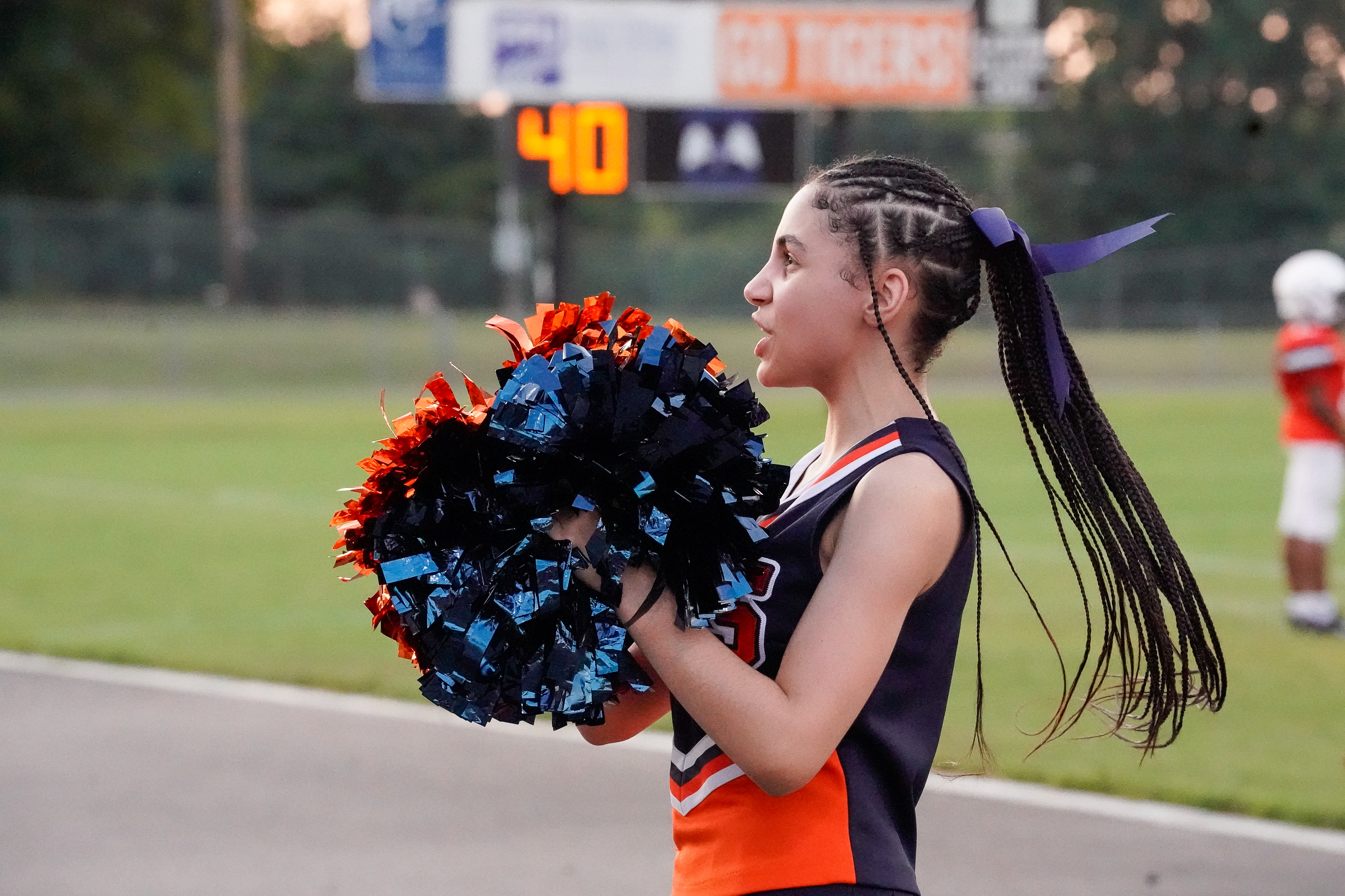 A young cheerleader with braided hair and pom-poms stands on a field with a scoreboard in the background.