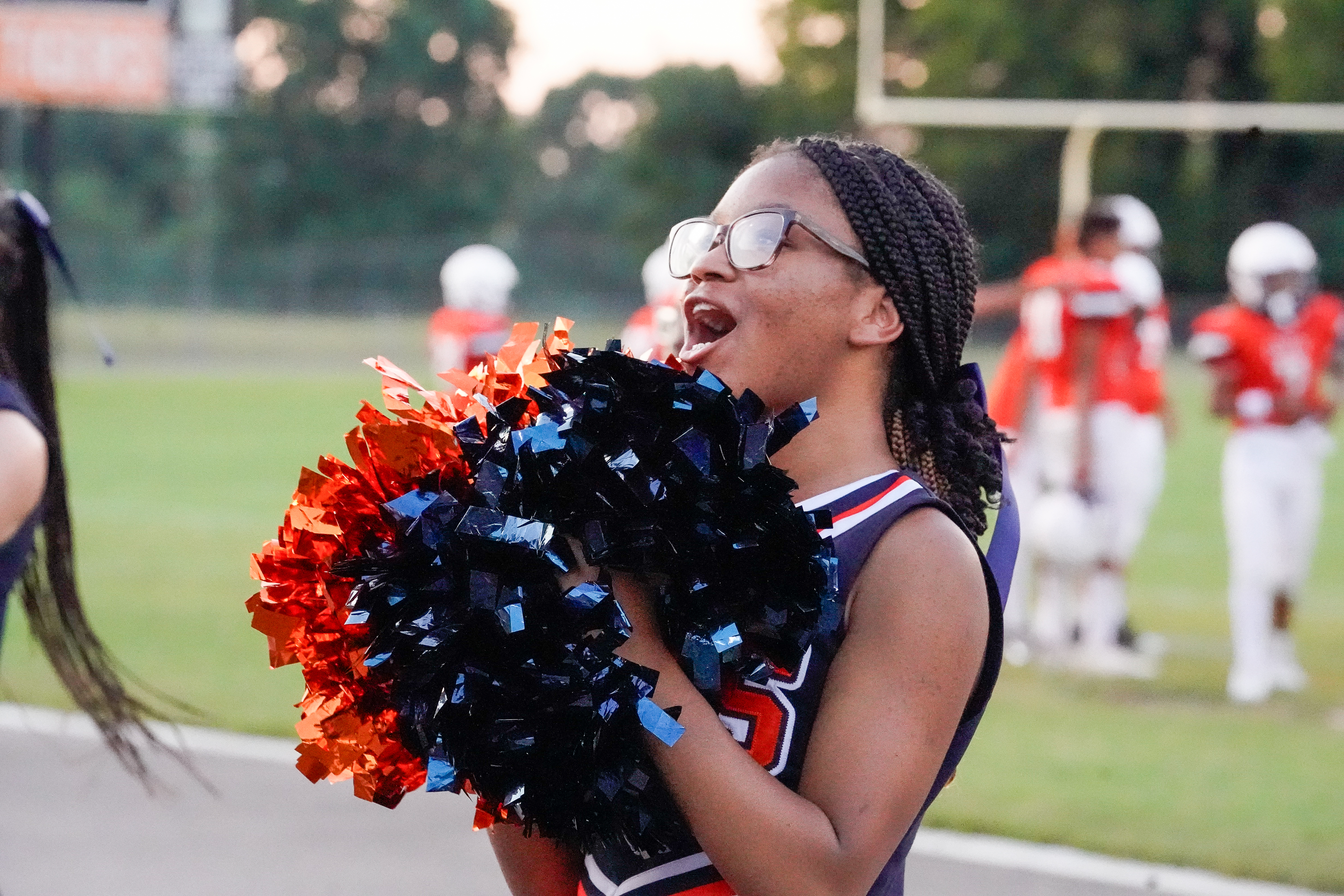 A young cheerleader with braided hair and glasses shouts enthusiastically while holding orange and navy pom-poms, with a blurred background of football players on a field.