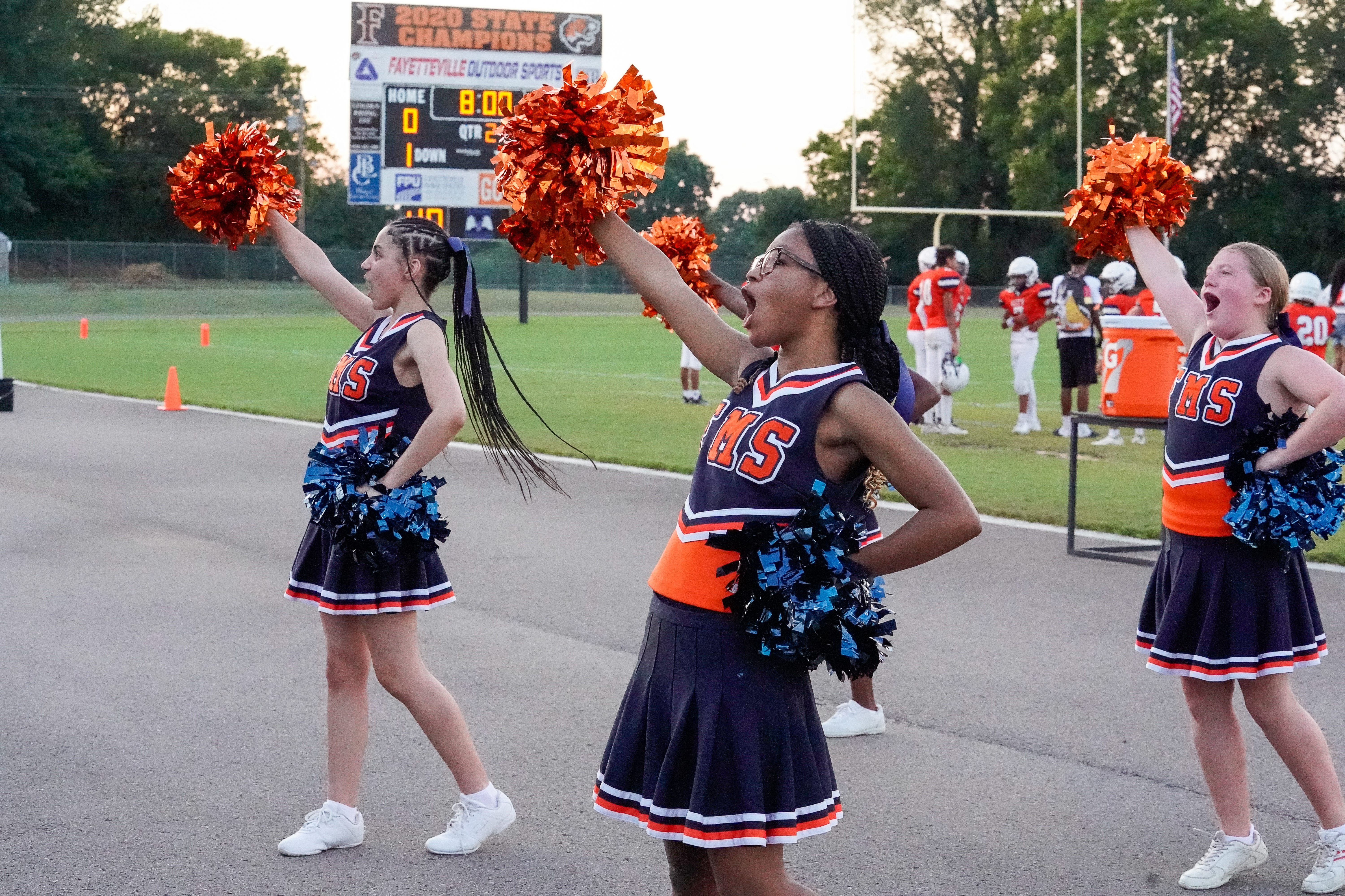 Three young cheerleaders in navy and orange uniforms with pom-poms are performing in the foreground, while a football field with players and a scoreboard is visible in the background.